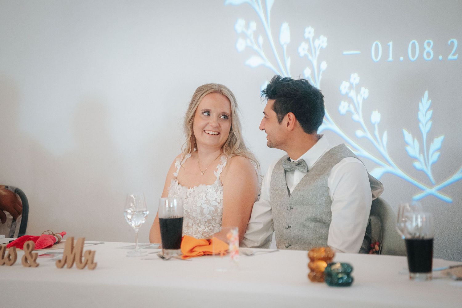 Smiling bride in a white lace dress and groom in a vest and bow tie sit at a wedding reception table with a floral projection behind them.