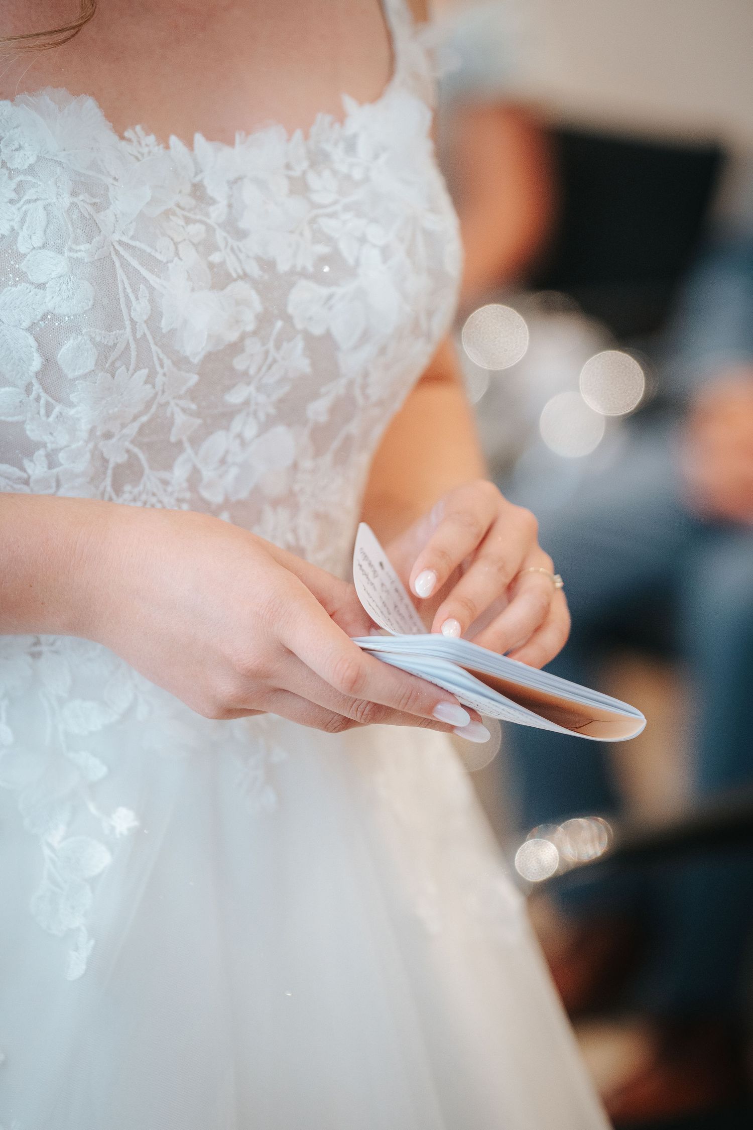 Close-up of a bride in an embroidered floral lace wedding gown holding small folded vow cards in her hands during the ceremony.