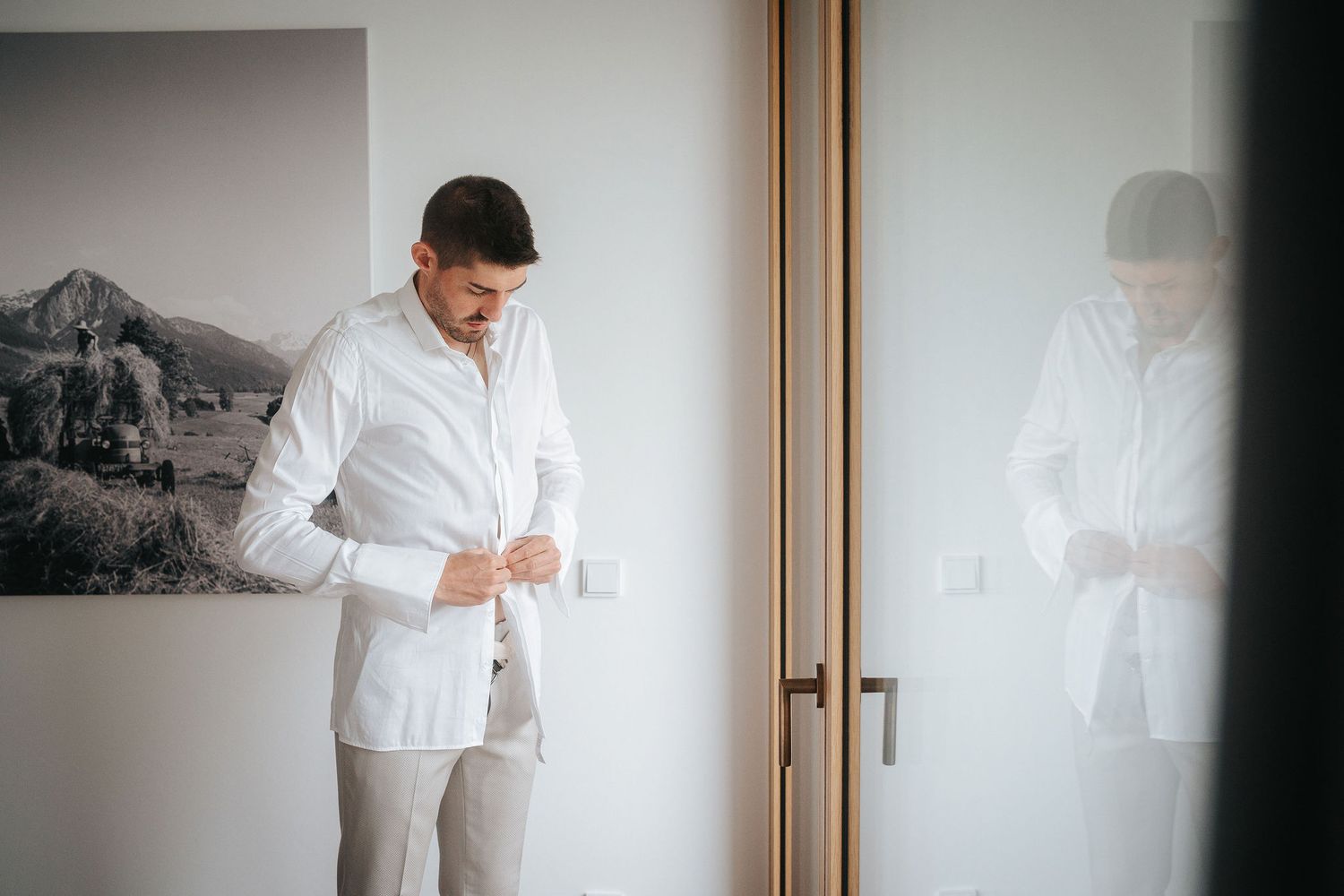 Groom in a white dress shirt and beige trousers buttons his cuffs while getting ready beside a mirrored door and a mountain landscape photo.