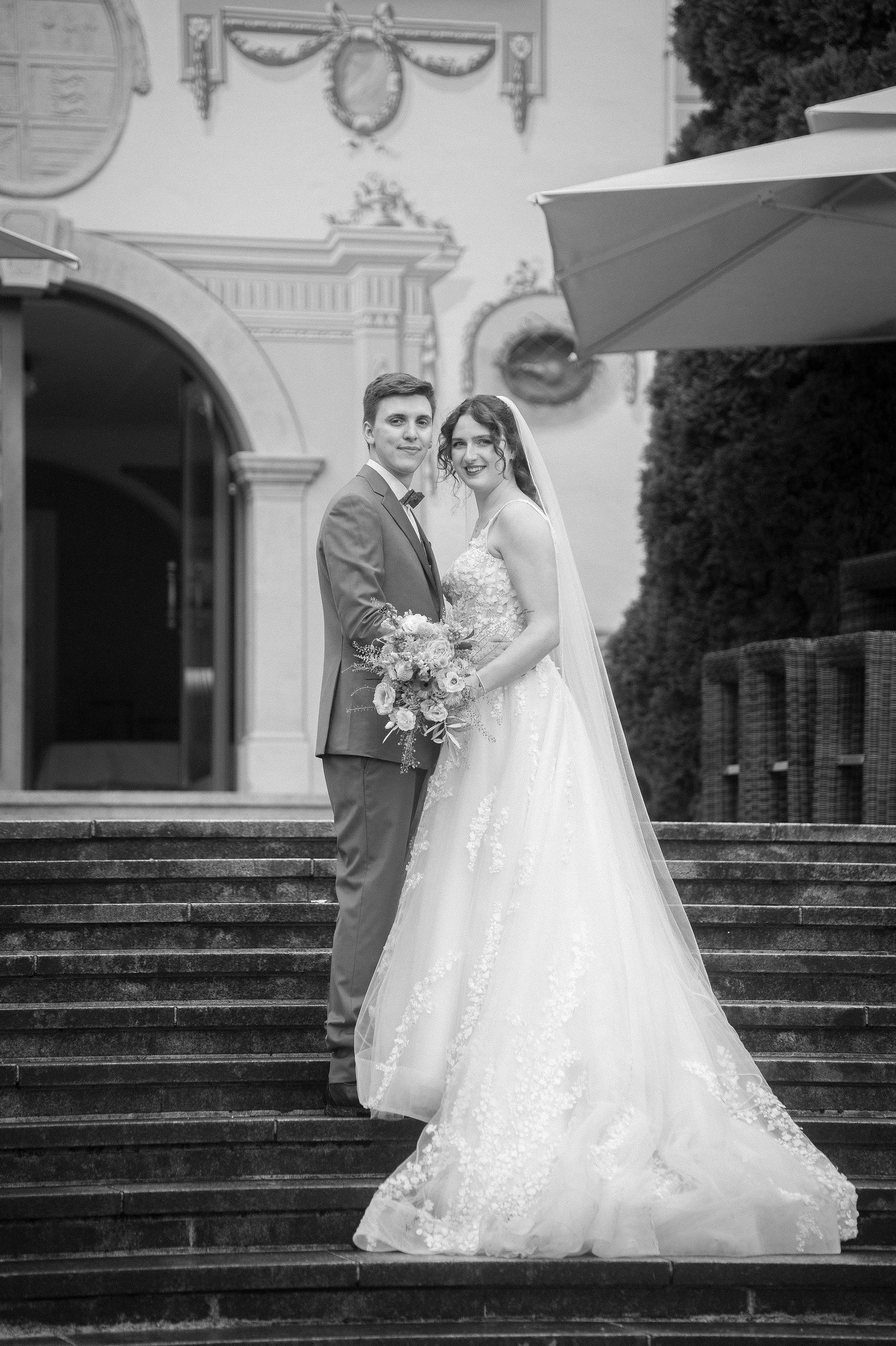Black and white photo of a bride in a lace gown with a long veil and a groom in a suit holding a bouquet on stone steps.