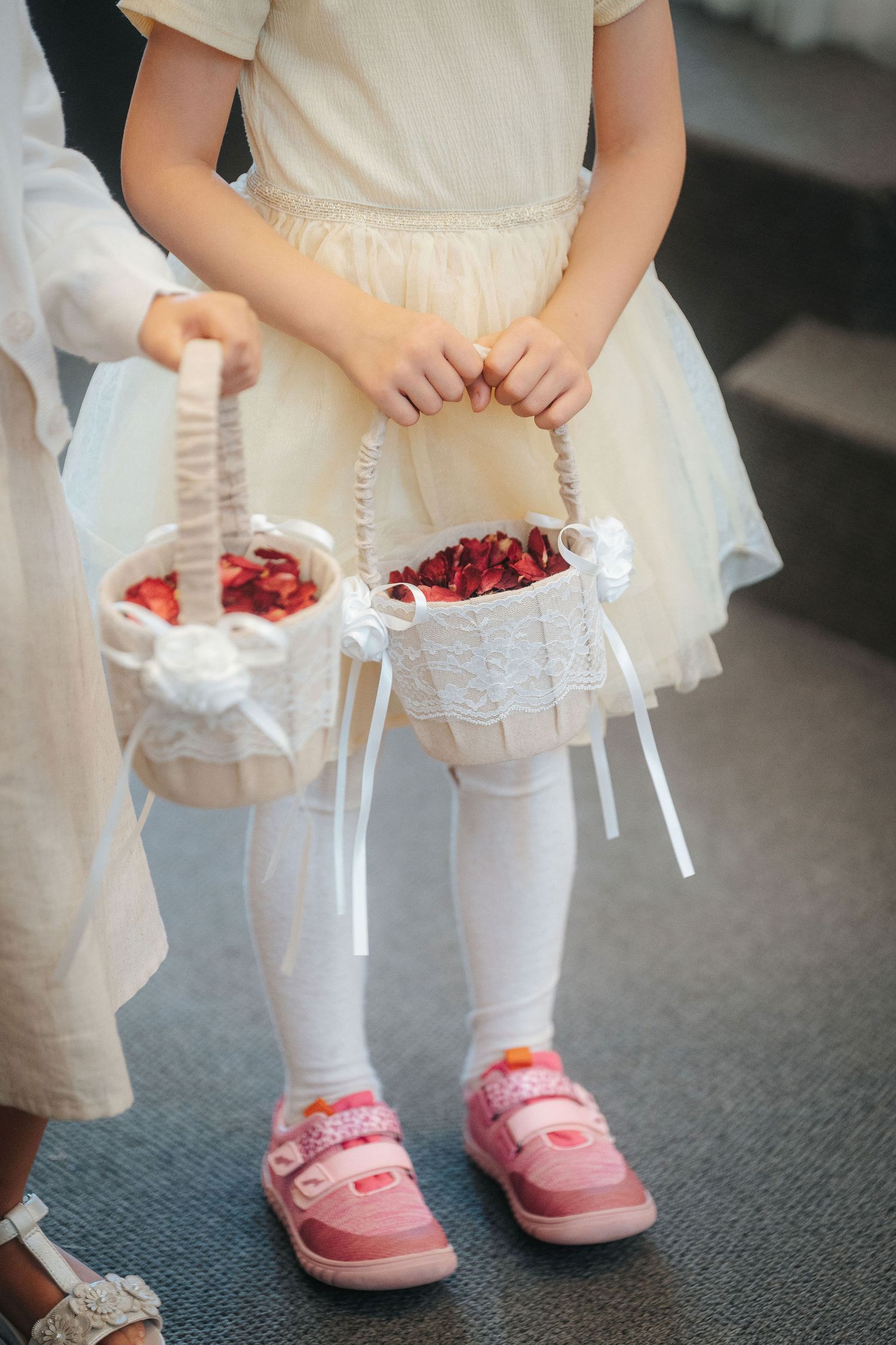 Flower girl in a cream tulle dress and pink sneakers holds a lace-trimmed basket filled with red rose petals at a wedding.
