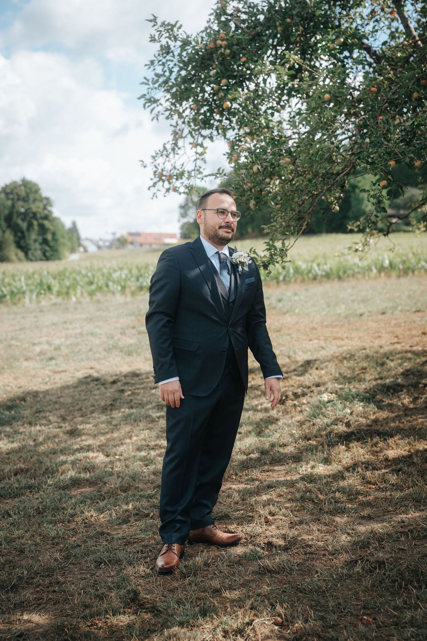Groom in a navy suit, tie and brown leather shoes stands under an apple tree in a sunlit grassy field, with a cornfield behind.