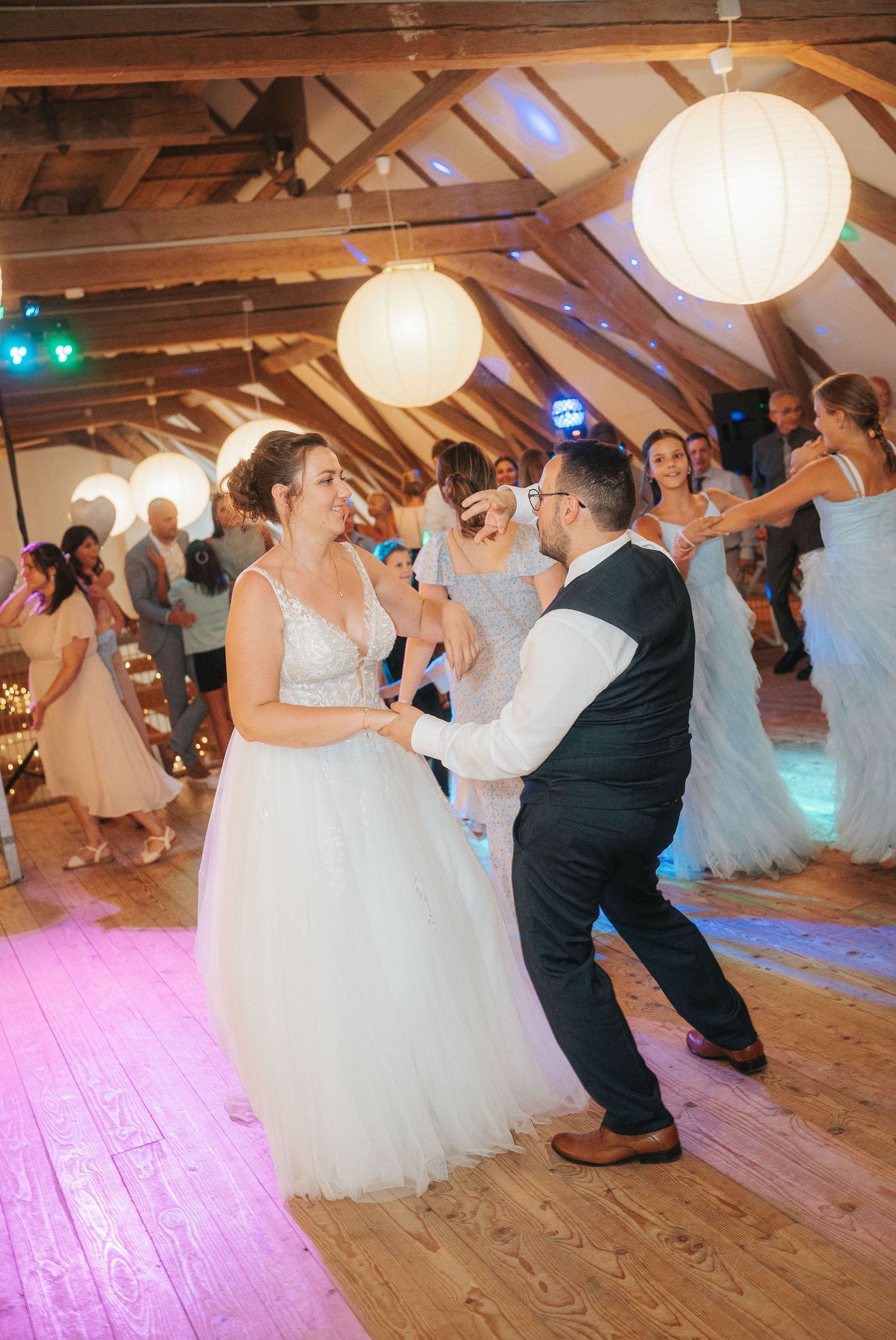 Bride in a white lace gown dances with a groom in a vest on a wooden floor at a rustic barn wedding reception with paper lanterns.