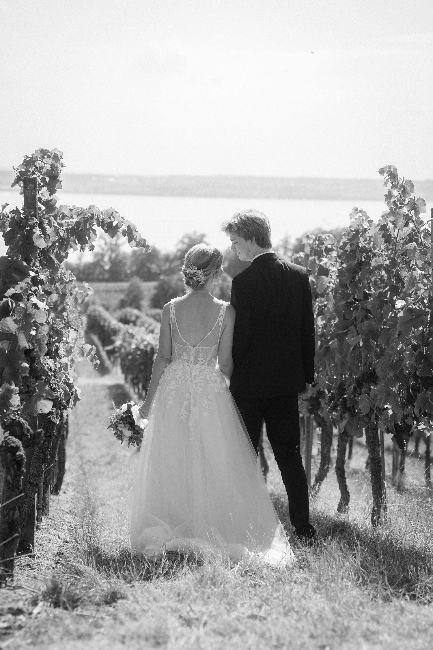 Black and white photo of a bride and groom walking hand in hand through a lush vineyard on their wedding day.