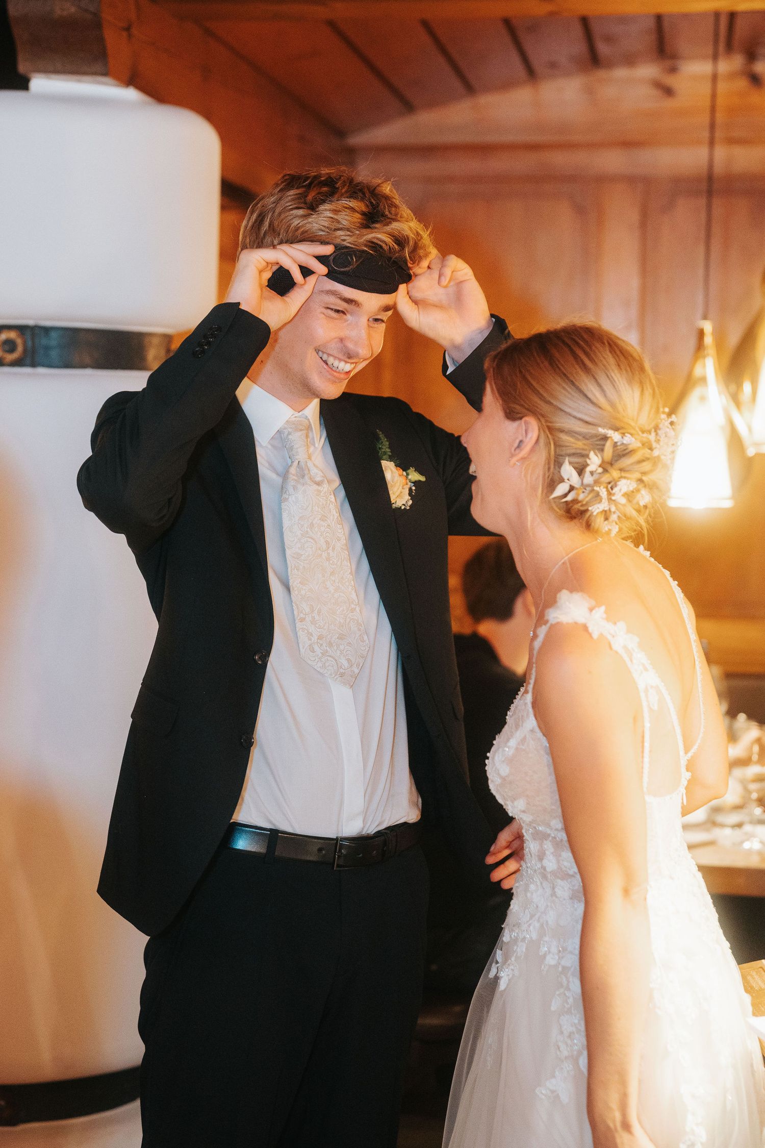 Smiling groom in a black suit adjusts a blindfold on his forehead while facing a bride in a lace gown with a floral hairpiece.
