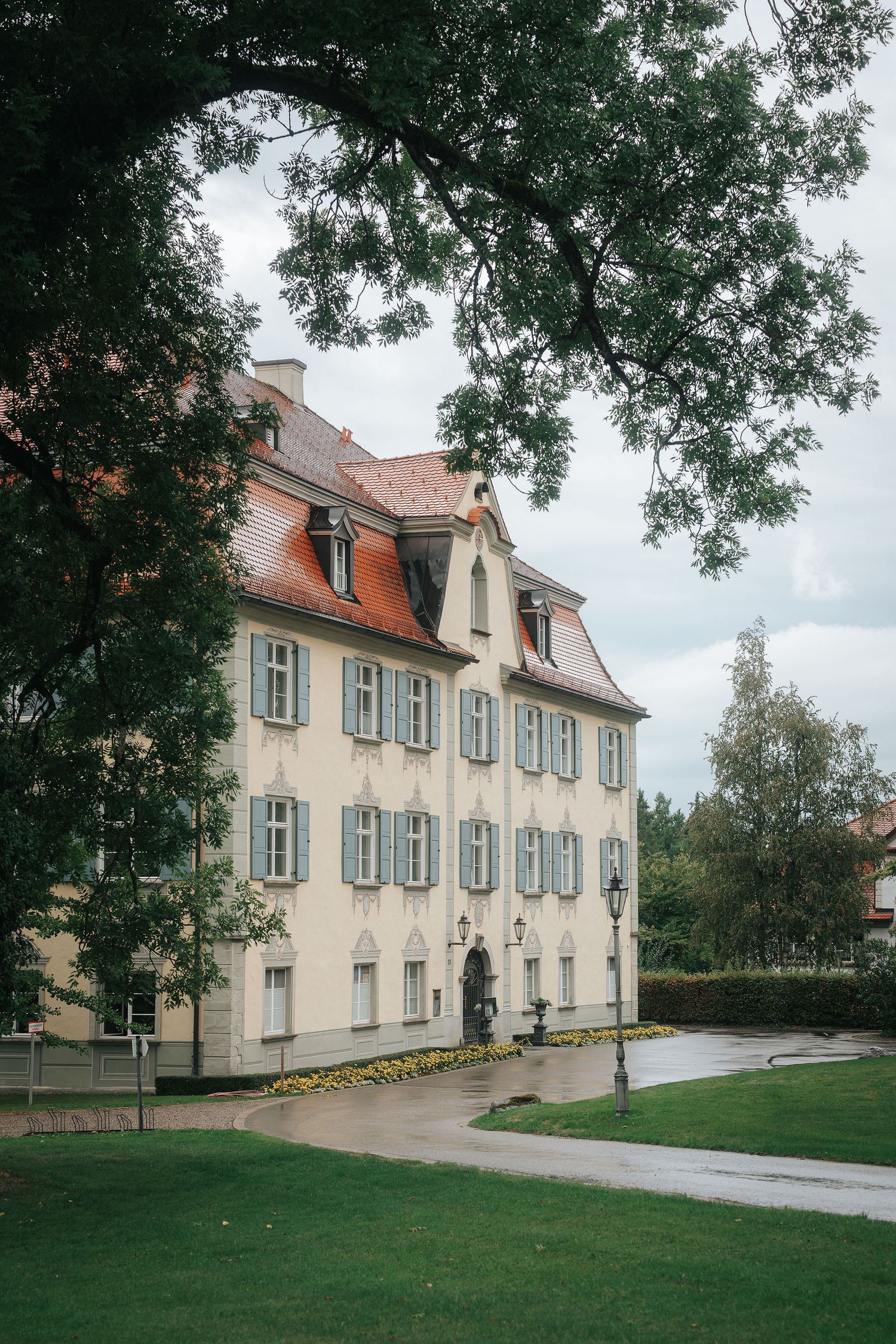 Cream-colored historic manor with red tile roof and teal shutters beside a curved wet path and green lawn on an overcast day.