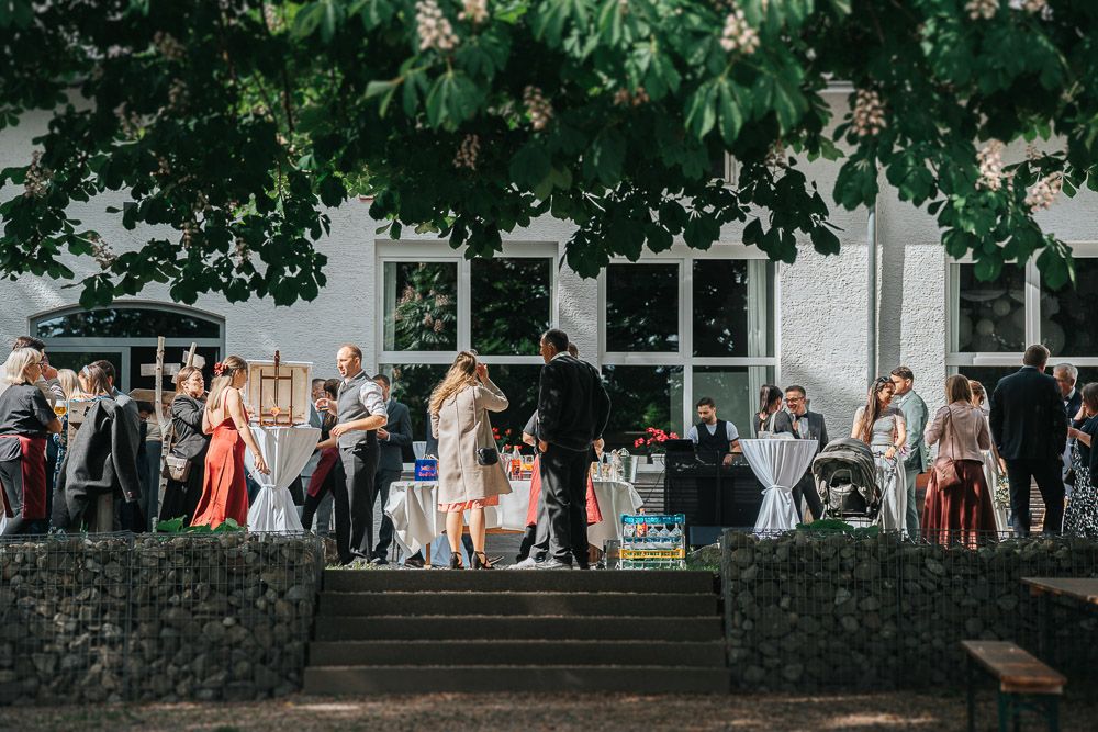 A group of people gather on stone steps outside a white building under leafy tree branches during a social event.