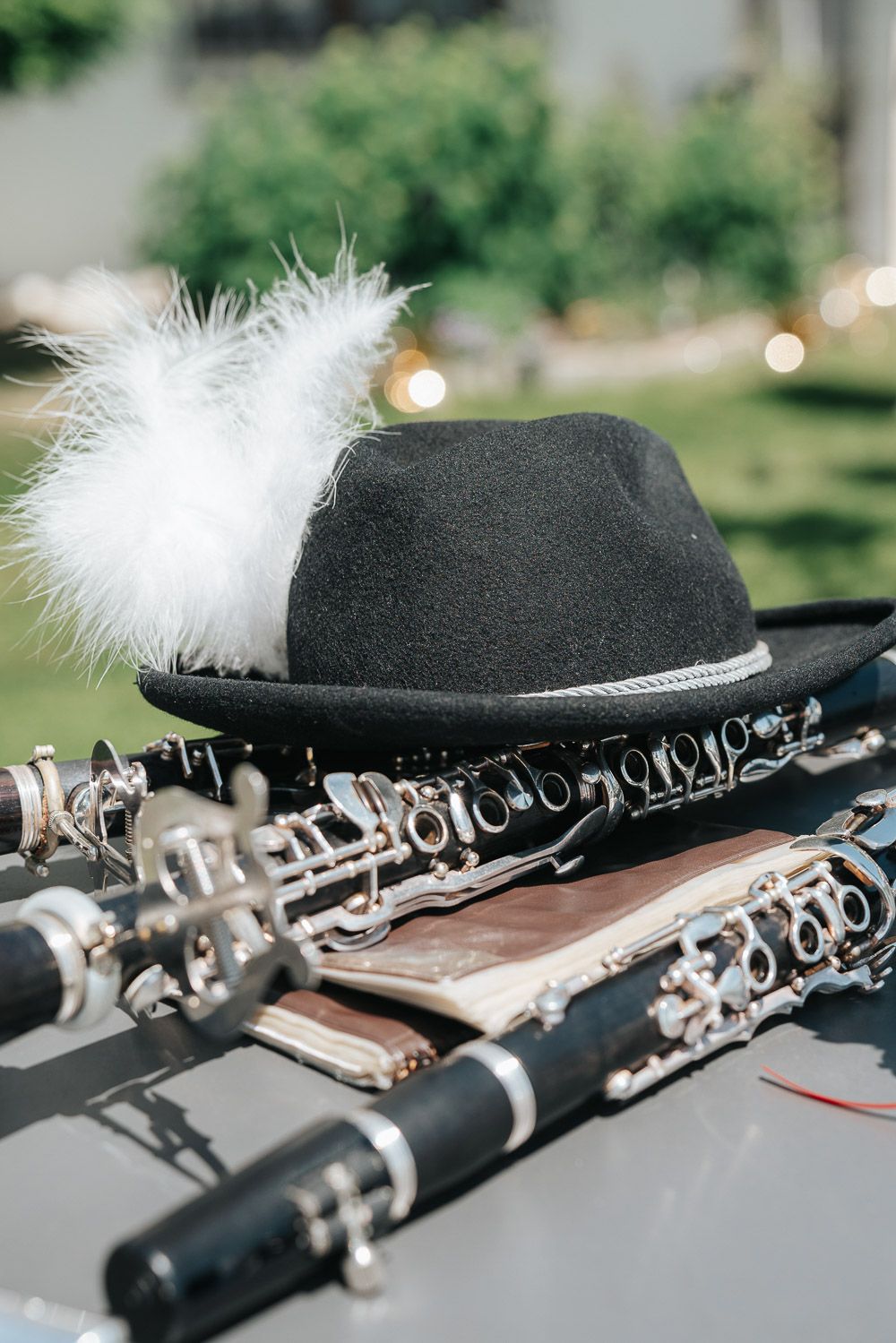 A black fedora hat with white feather sits atop a pair of wooden clarinets against a blurred outdoor background.
