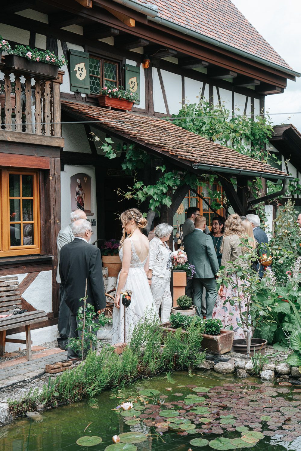Guests gather at a traditional rustic cottage venue with lily ponds and timber framing for an outdoor wedding celebration.