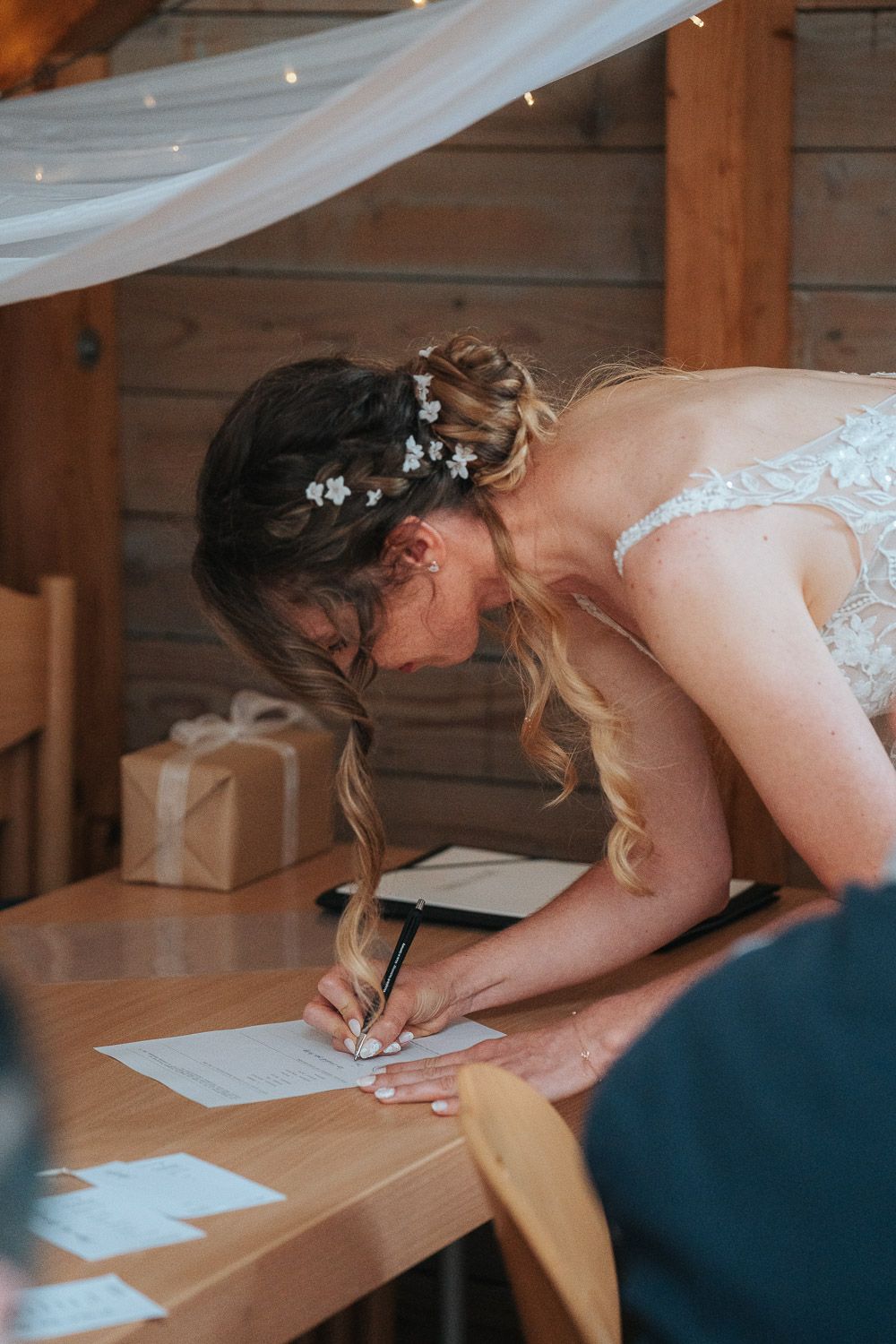 Someone in a white wedding dress leans over a wooden table to sign papers at an indoor venue with sheer drapery.