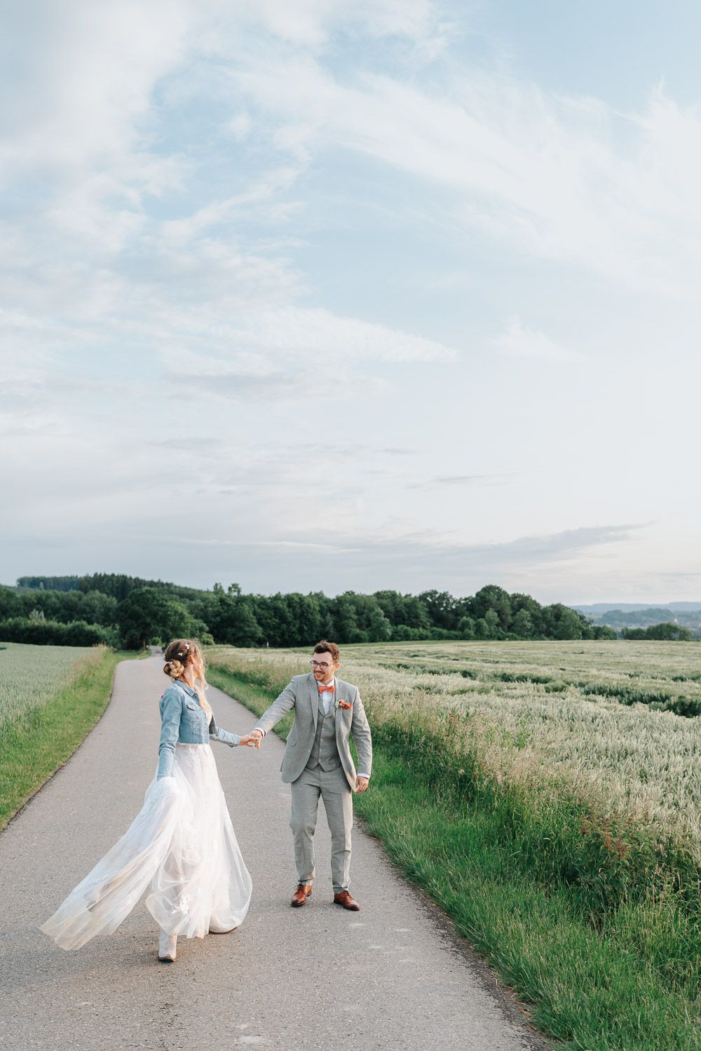 A couple walks hand in hand on a country path surrounded by lush green fields and a soft cloudy sky.