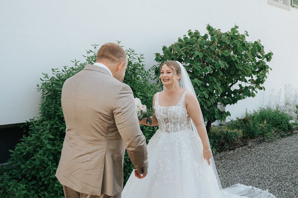A couple shares their first look moment on their wedding day beside lush greenery near a white wall.