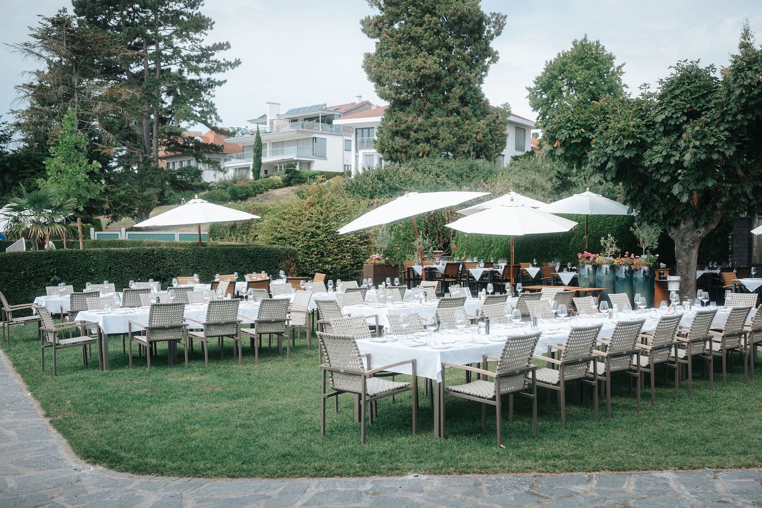 Long outdoor wedding reception tables with white linens and chairs on a green lawn beneath white parasols and trees.