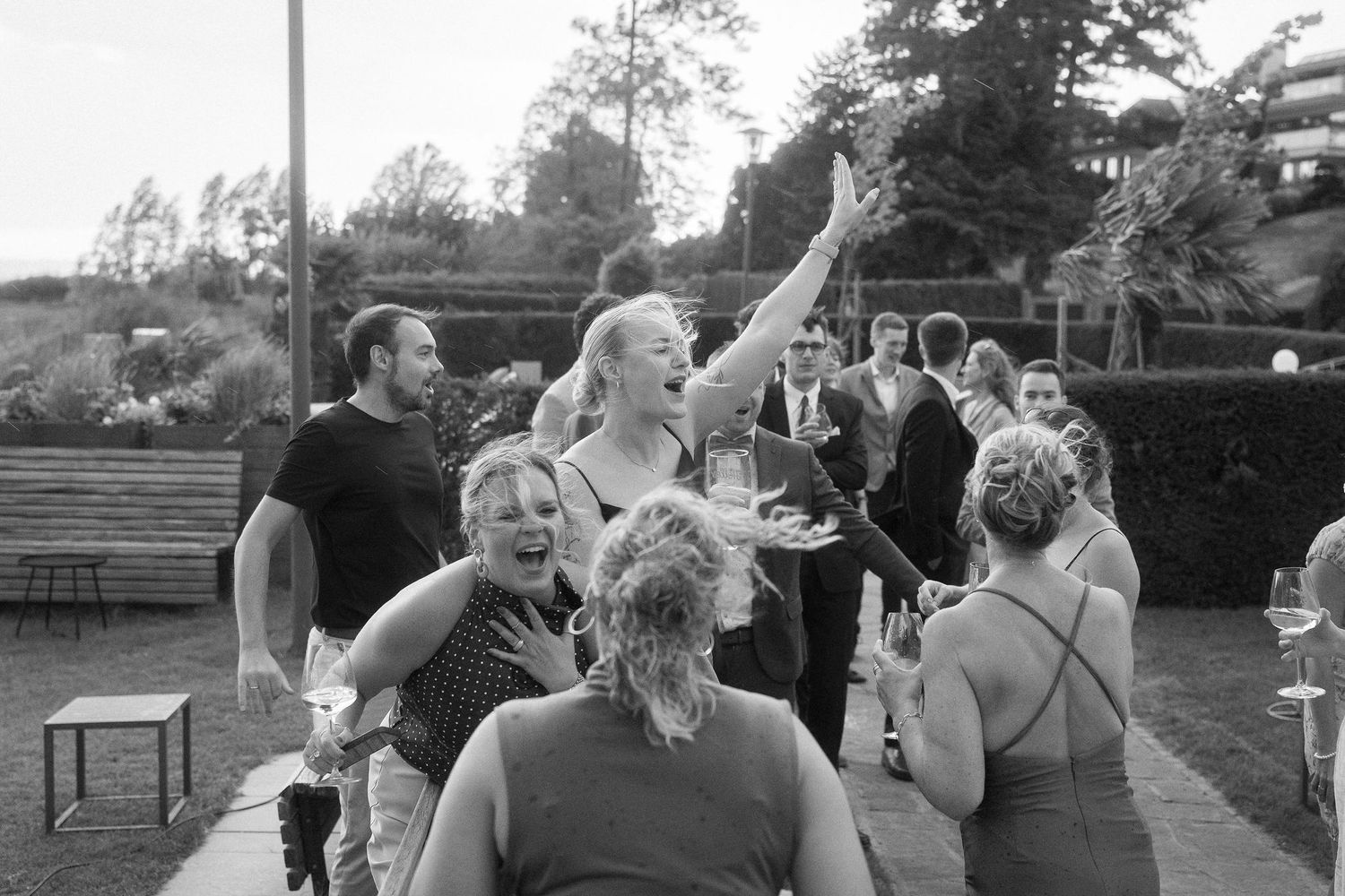 Black and white photo of joyful wedding guests laughing and cheering with raised arms and drinks at an outdoor garden celebration.