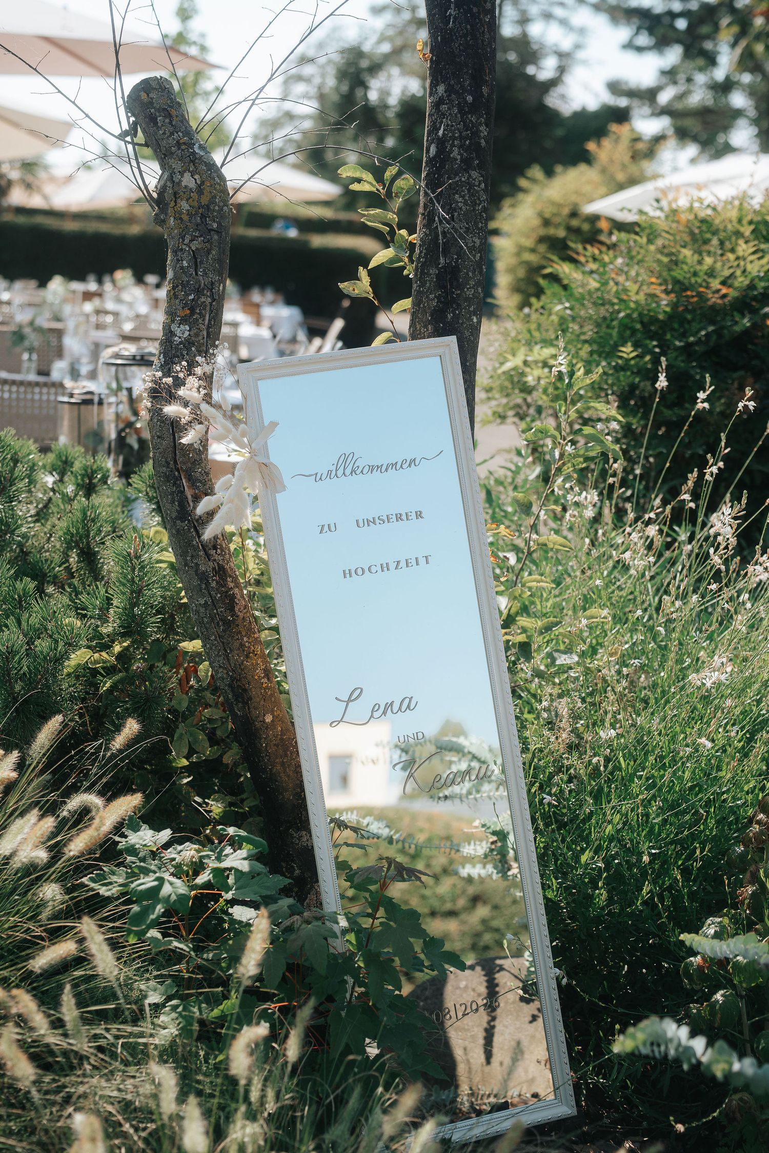 Wedding welcome mirror sign reading Willkommen zu unserer Hochzeit, tied to a tree among tall grasses with an outdoor reception area behind.