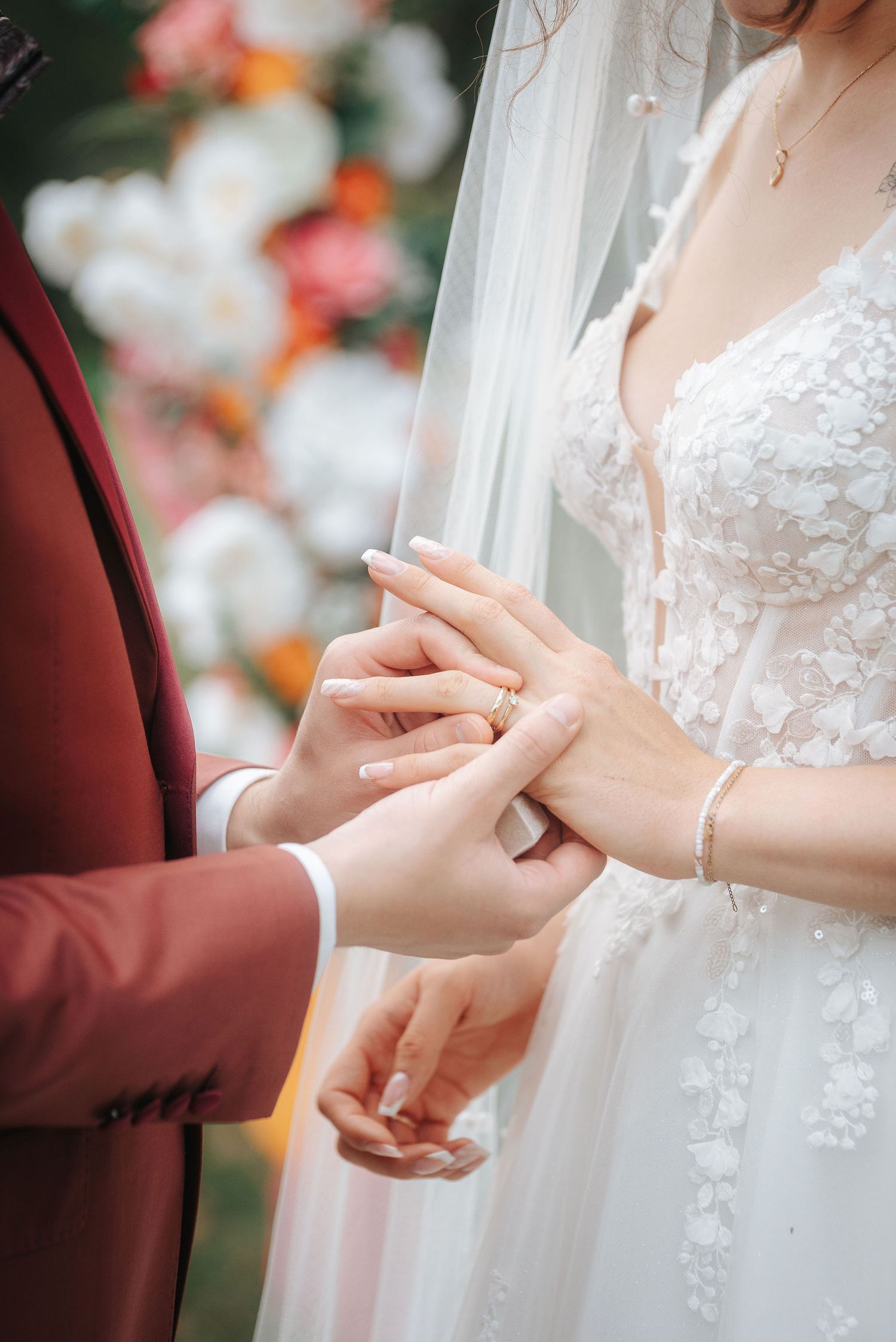 Groom in a burgundy suit slides a gold wedding ring onto the bride's finger during an outdoor ceremony with floral decor in the background.