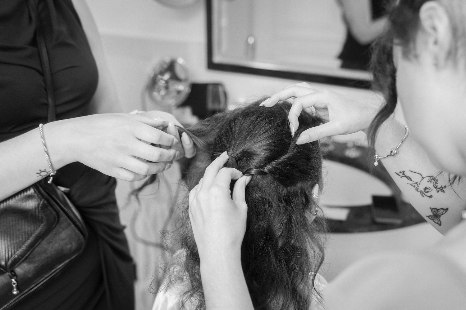Black and white photo of two hairstylists braiding and pinning a woman's long wavy hair in a bright salon.