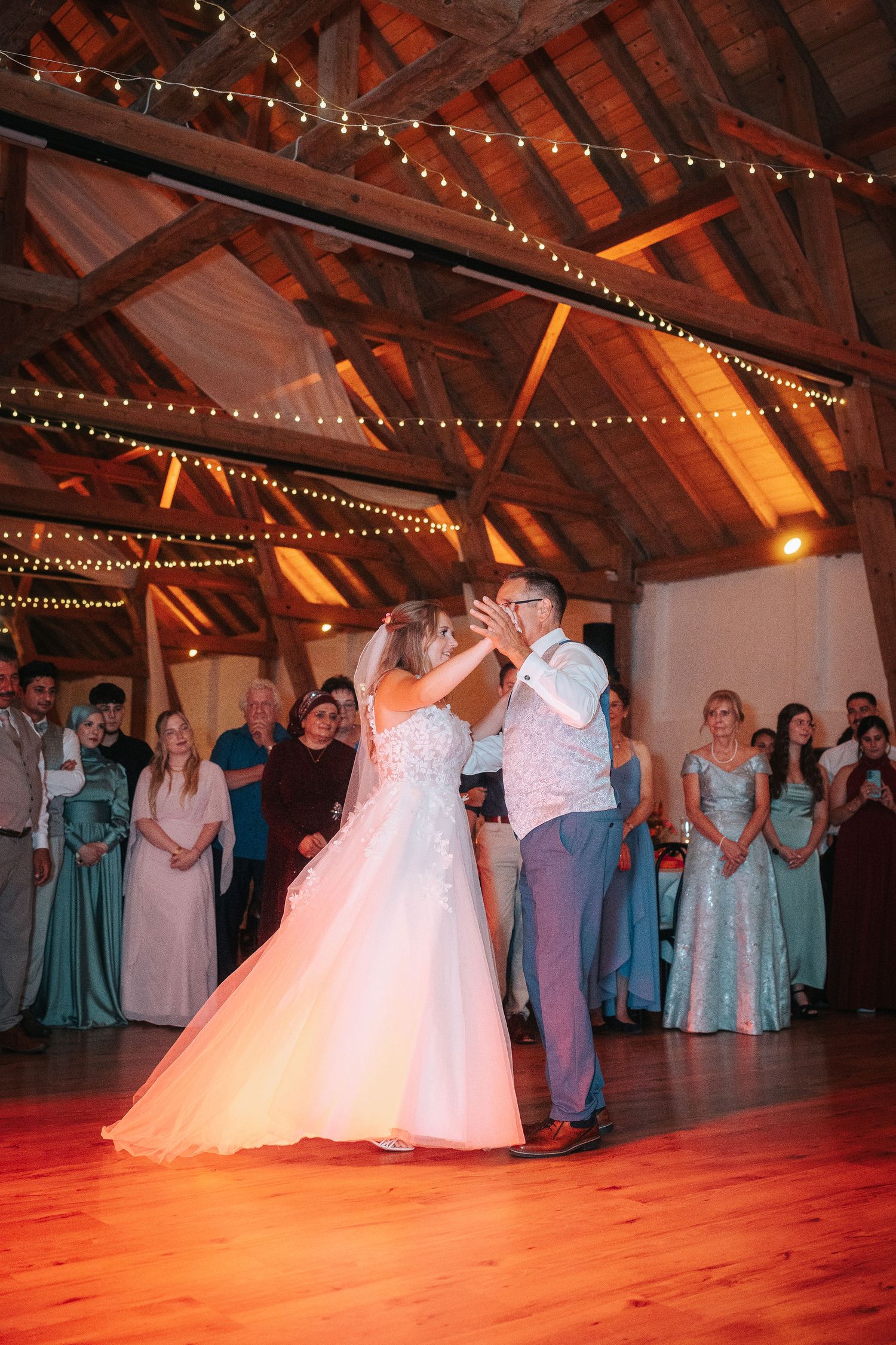 Bride in a white gown and groom share a first dance on a wooden floor as wedding guests watch inside a rustic barn venue.