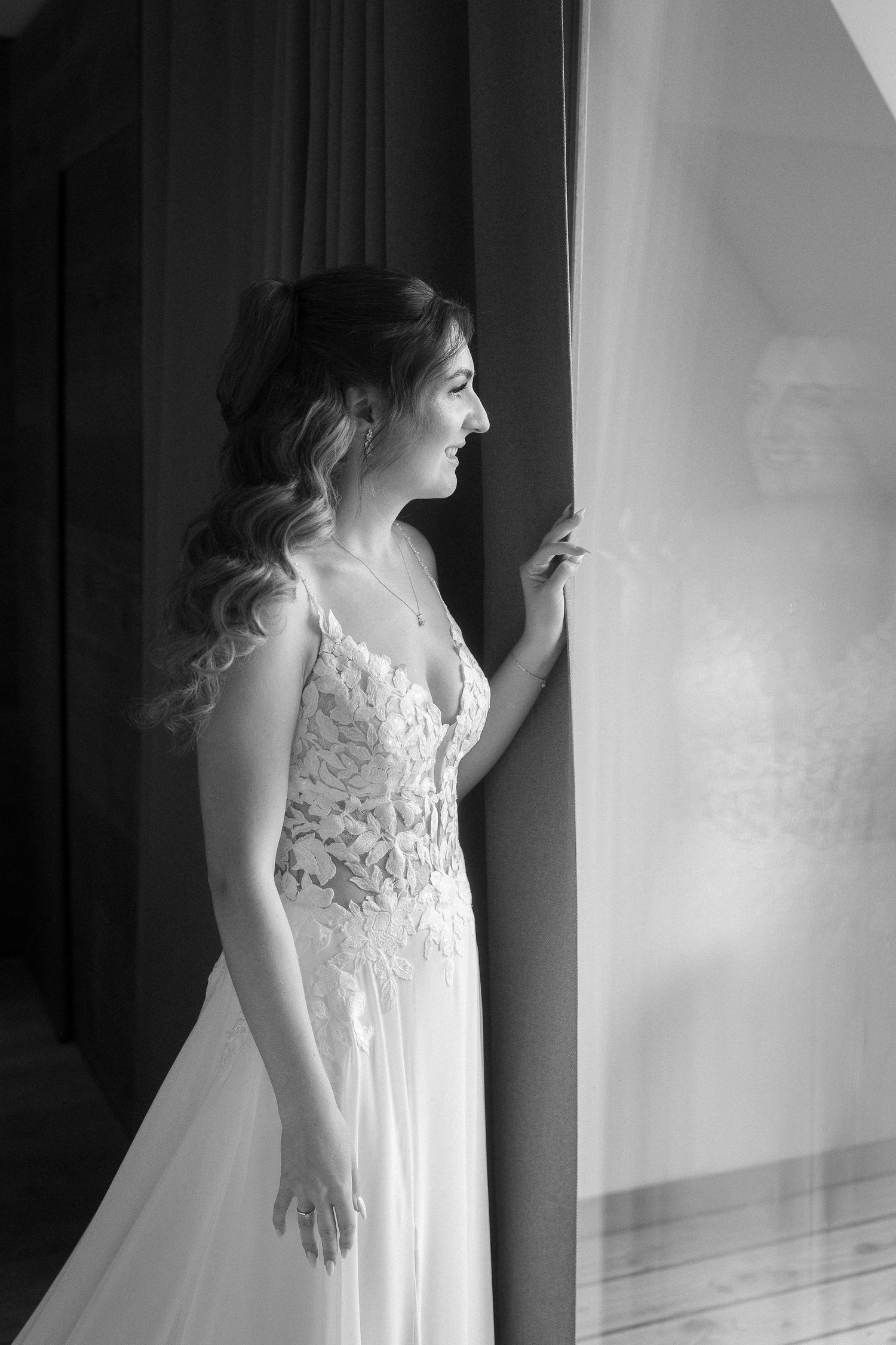 Black and white photo of a bride in a lace wedding gown with wavy hair gazing out a window beside a sheer curtain.