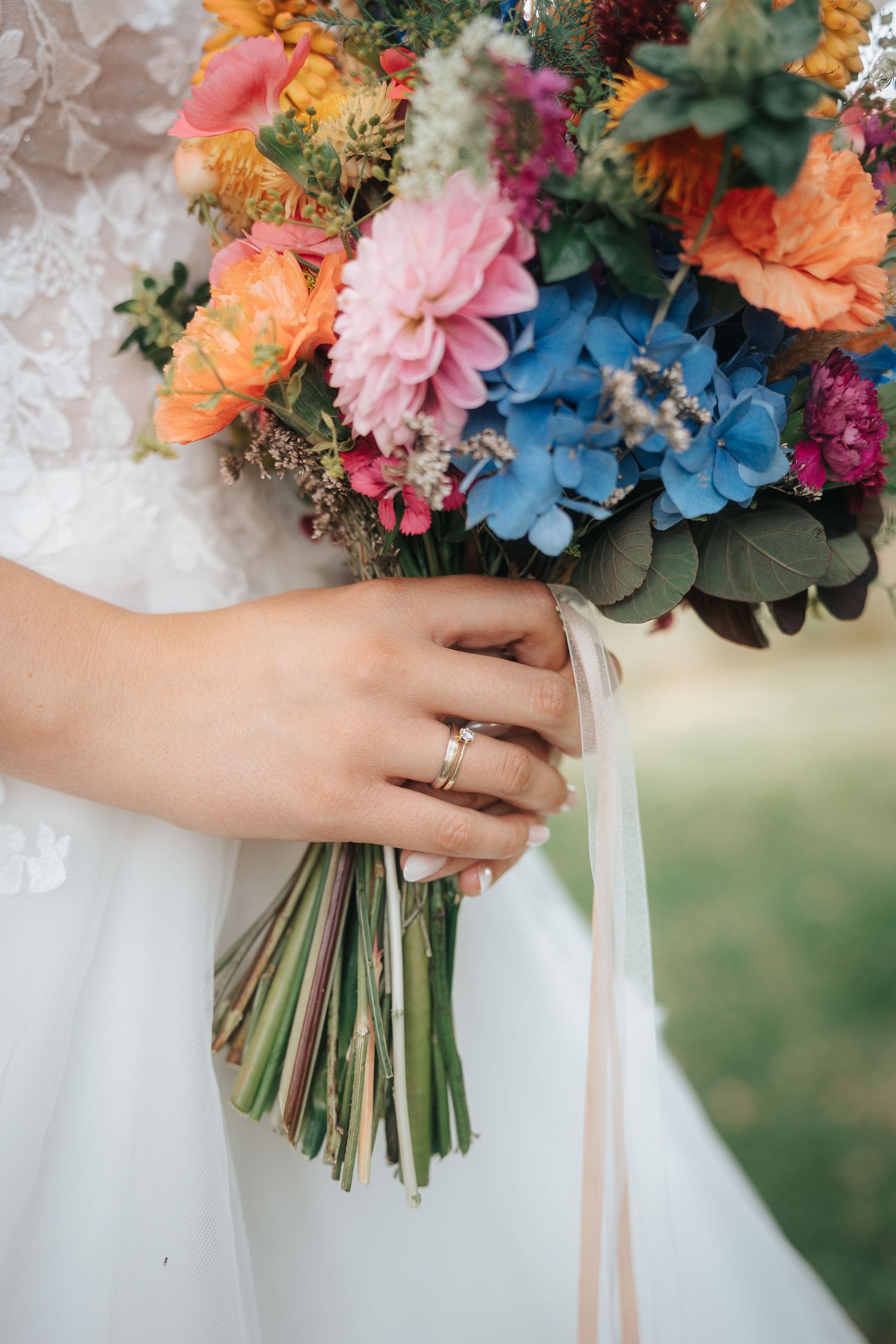 Bride in a lace wedding dress holds a colorful bouquet of pink, orange and blue hydrangea flowers with ribbon-wrapped stems.