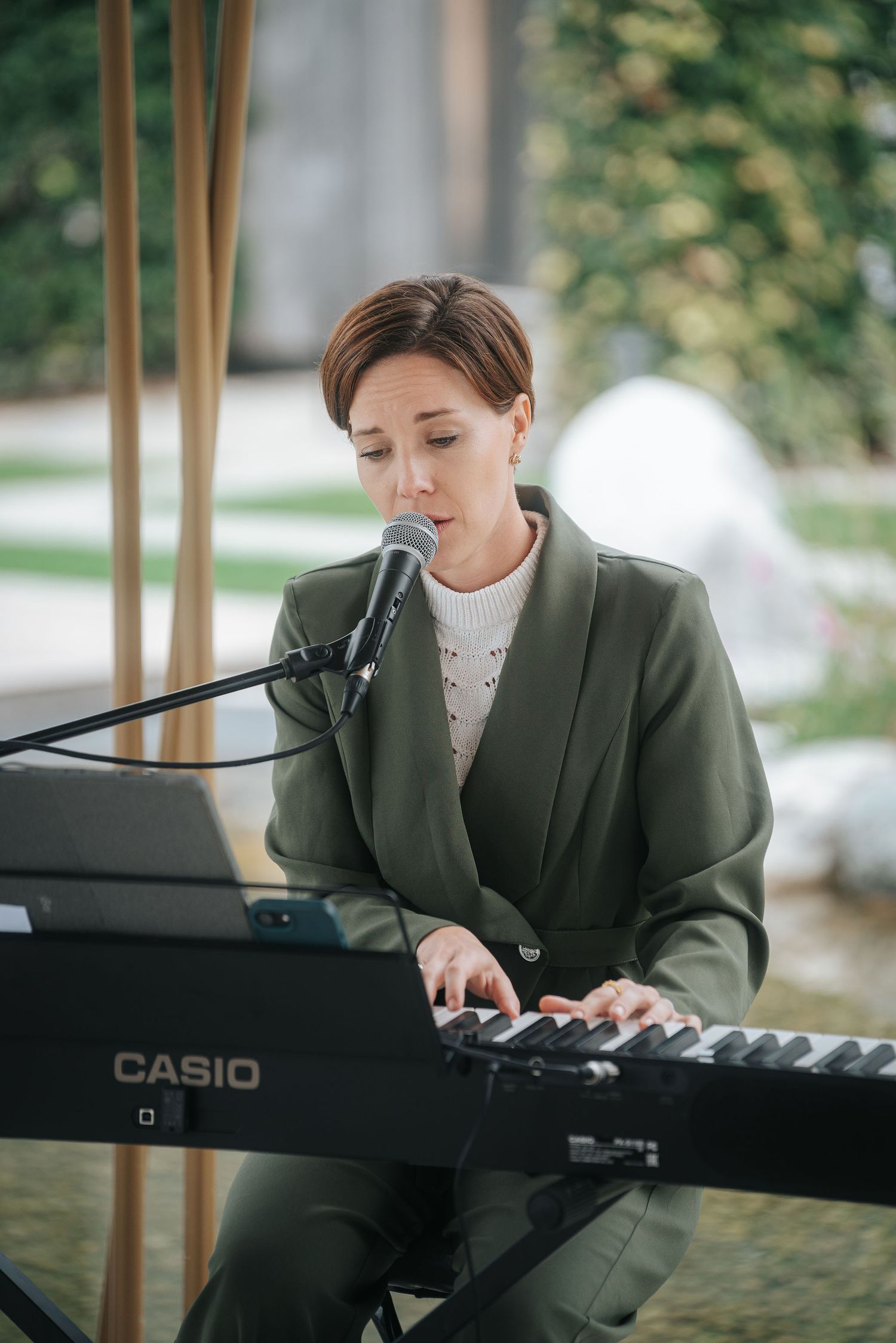 Woman in a green blazer sings into a microphone while playing a black Casio keyboard at an outdoor event with greenery behind her.