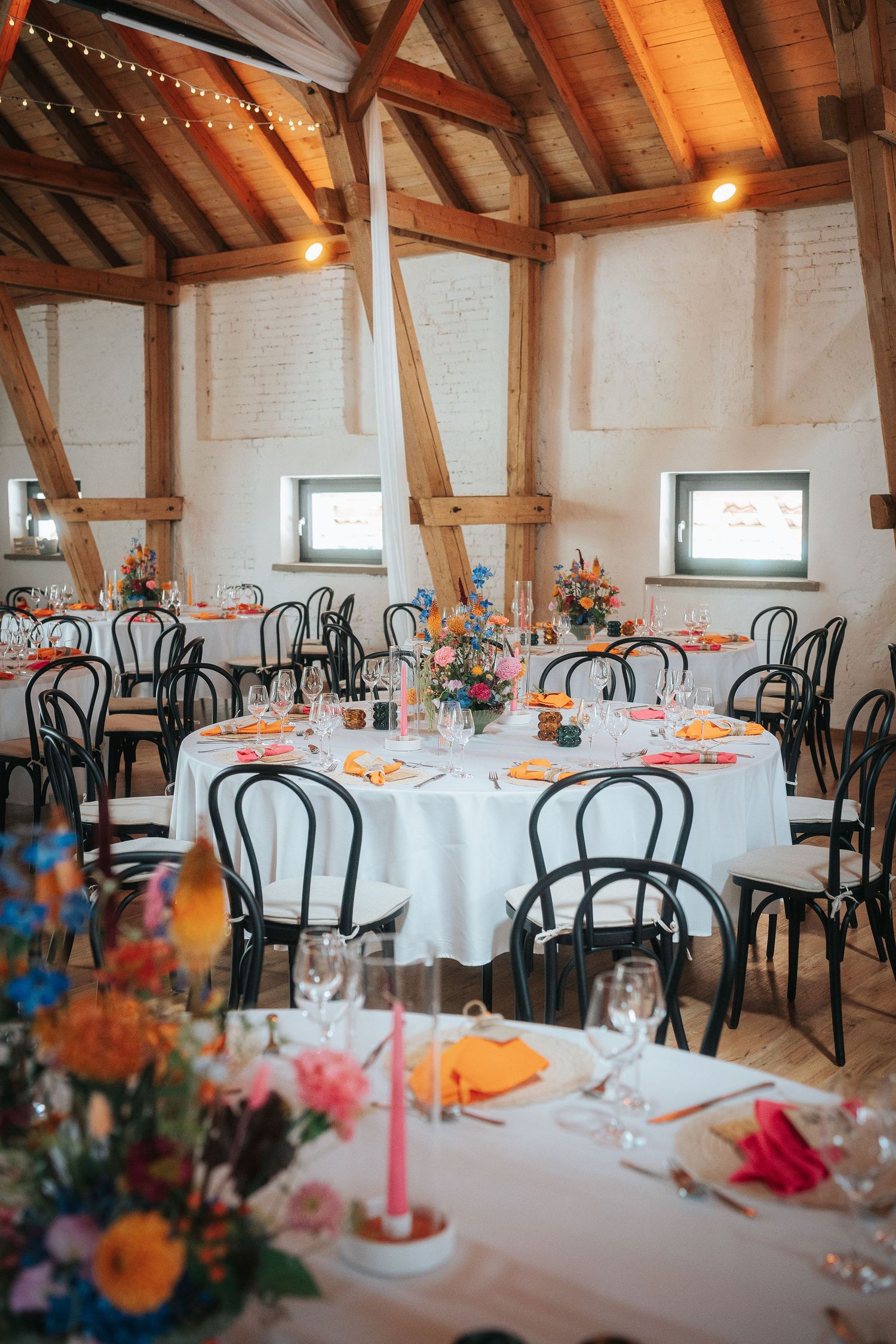 Rustic barn wedding reception with round tables, black chairs, colorful floral centerpieces and exposed timber beams.