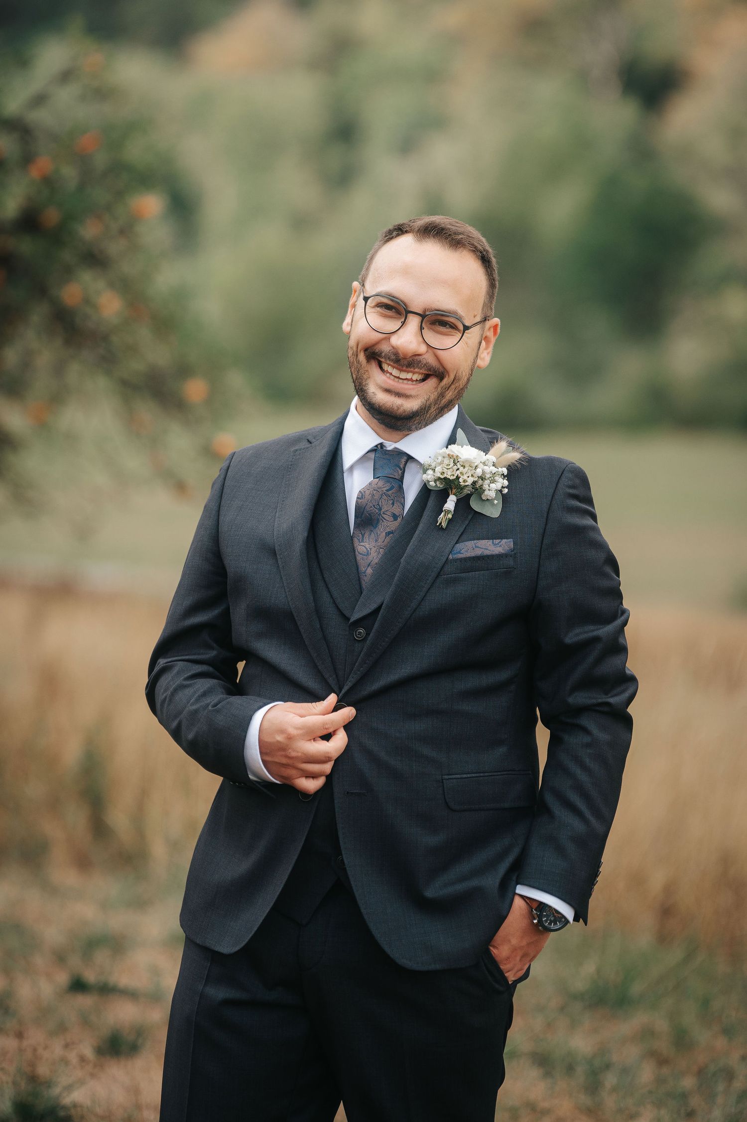 Smiling groom in a dark three-piece suit with glasses and a floral boutonniere poses outdoors in a golden autumn field.
