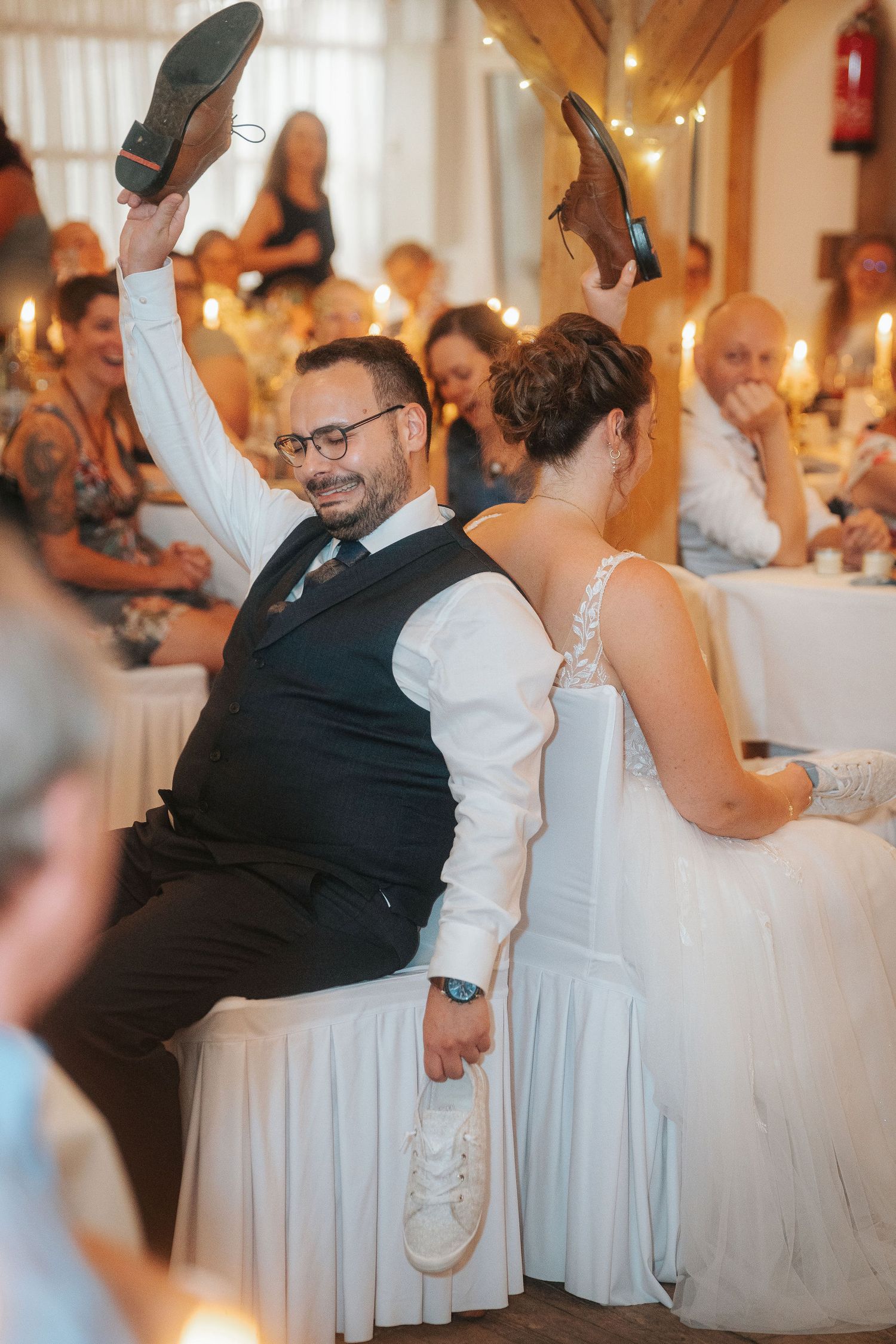 Bride and groom sit back to back holding up shoes during a lively wedding reception shoe game as guests laugh nearby.