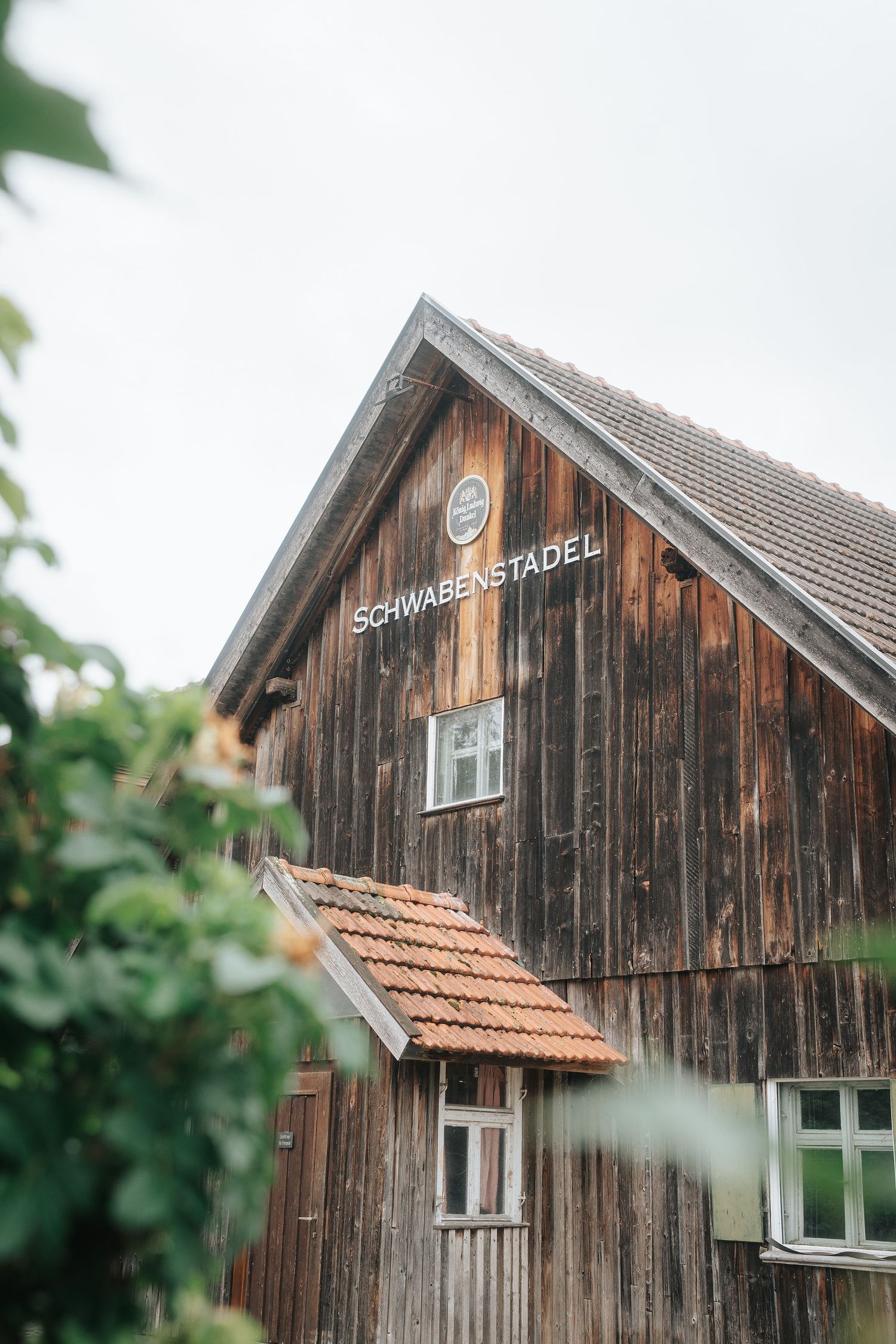 Weathered wooden barn with a tiled roof and the sign Schwabenstadel, framed by green leaves under a soft overcast sky.