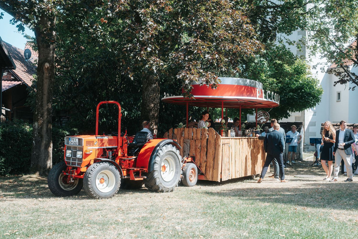 Vintage red tractor pulling a wooden hay wagon with a red canopy while visitors gather on a grassy lawn under shady trees.