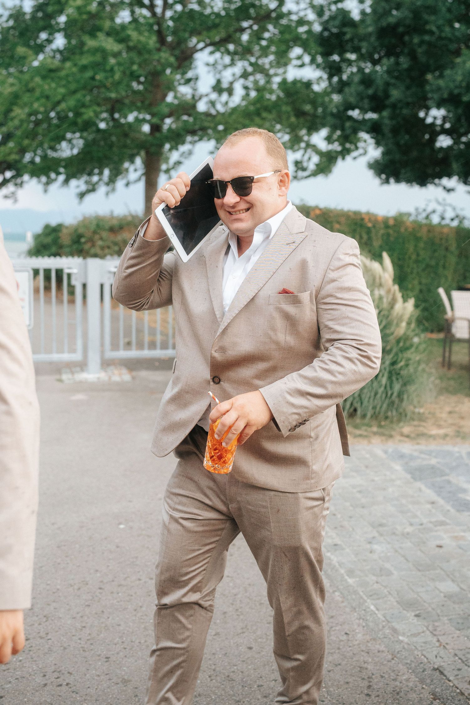 Man in sunglasses and a tan suit smiles while holding a tablet to his head and an orange drink outdoors near trees and a white fence.