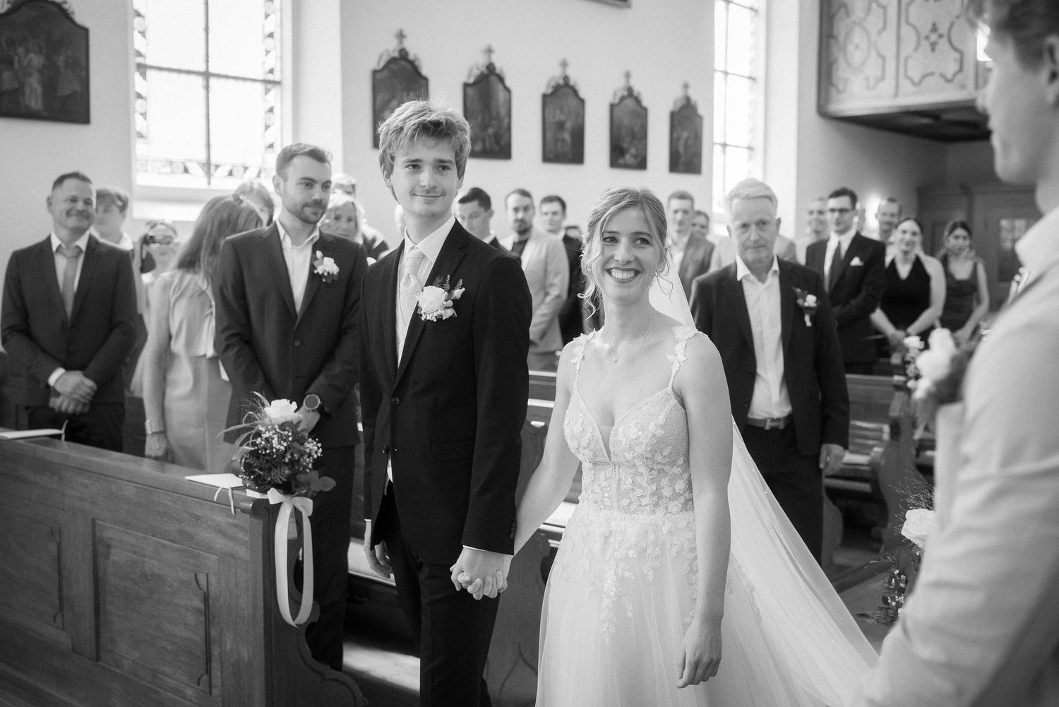 Smiling bride in a lace wedding gown holds hands with the groom at the church altar as wedding guests look on.