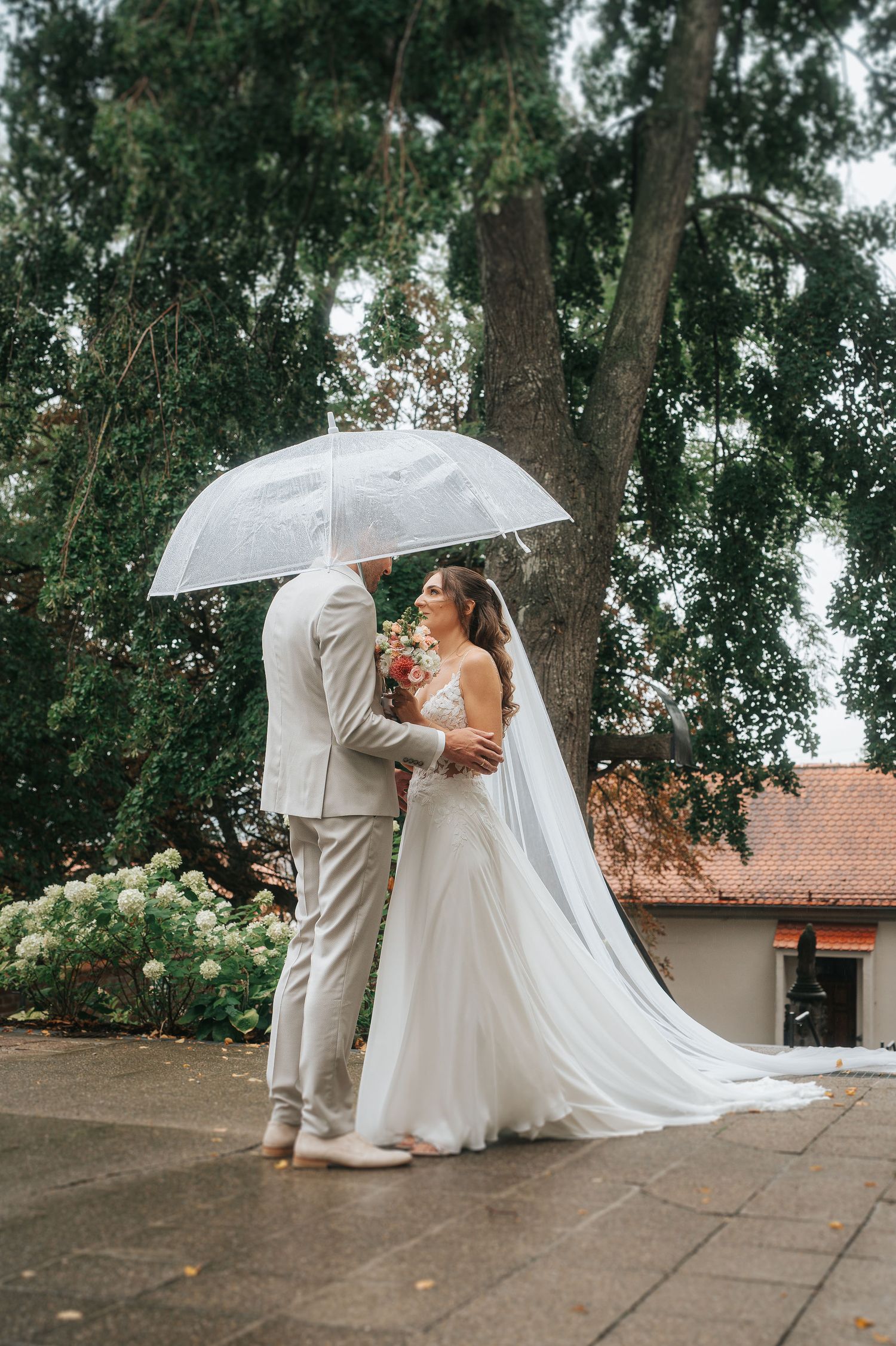 Bride in a flowing white gown and long veil embraces her groom in a light suit under a clear umbrella on a rainy wedding day outdoors.