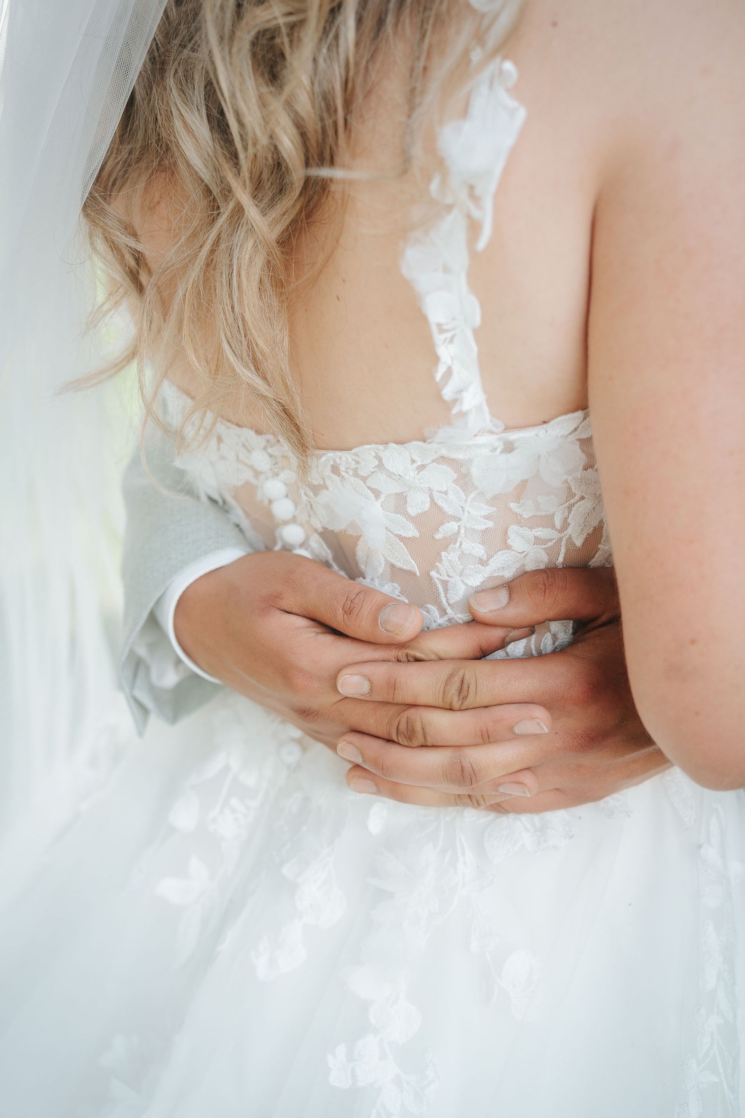 Groom's hands embrace a bride from behind, her lace wedding gown with floral appliqué and soft veil glowing in natural light.
