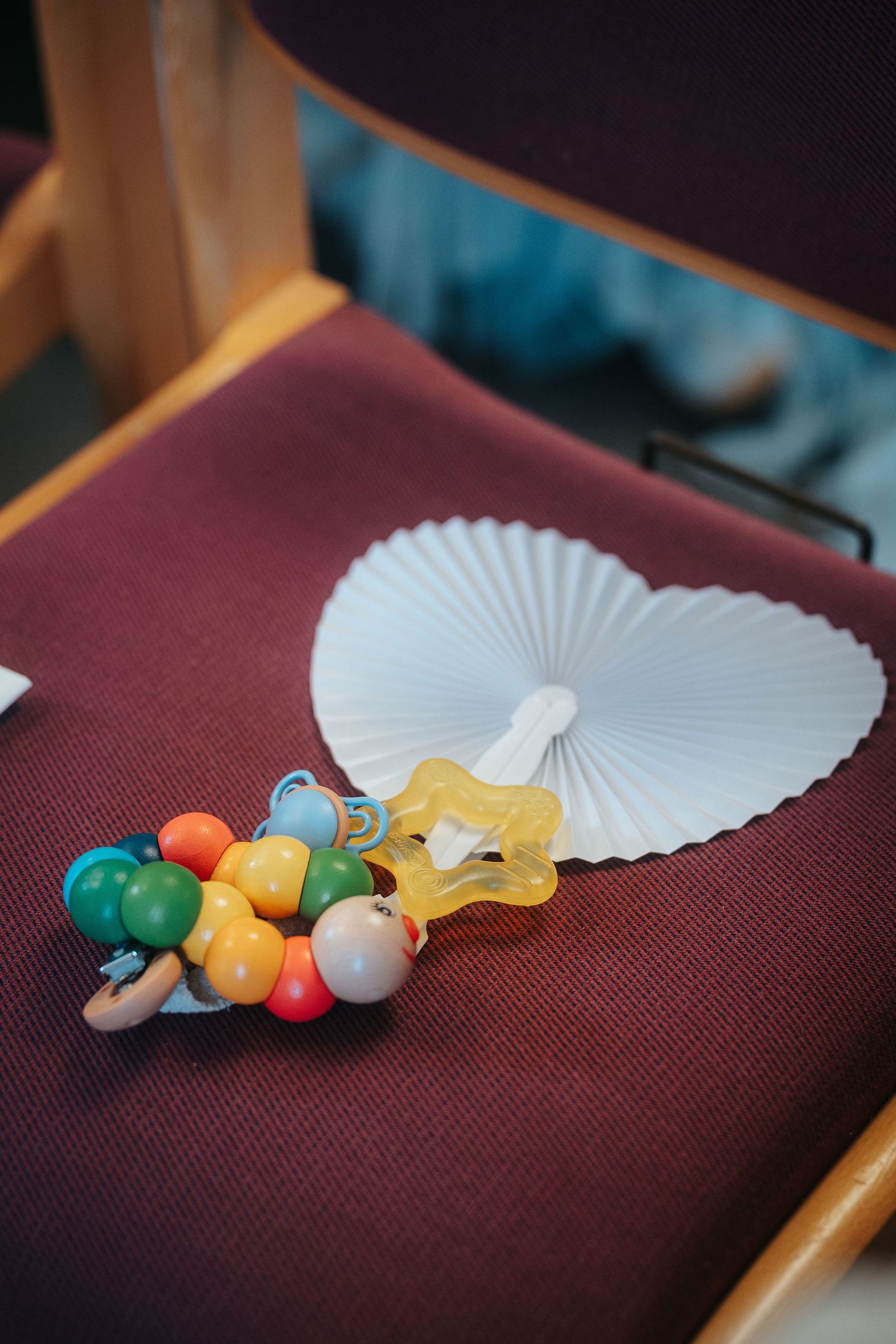 Colorful wooden bead baby rattle and yellow pacifier with a white heart-shaped paper fan resting on a burgundy cushioned wooden chair.