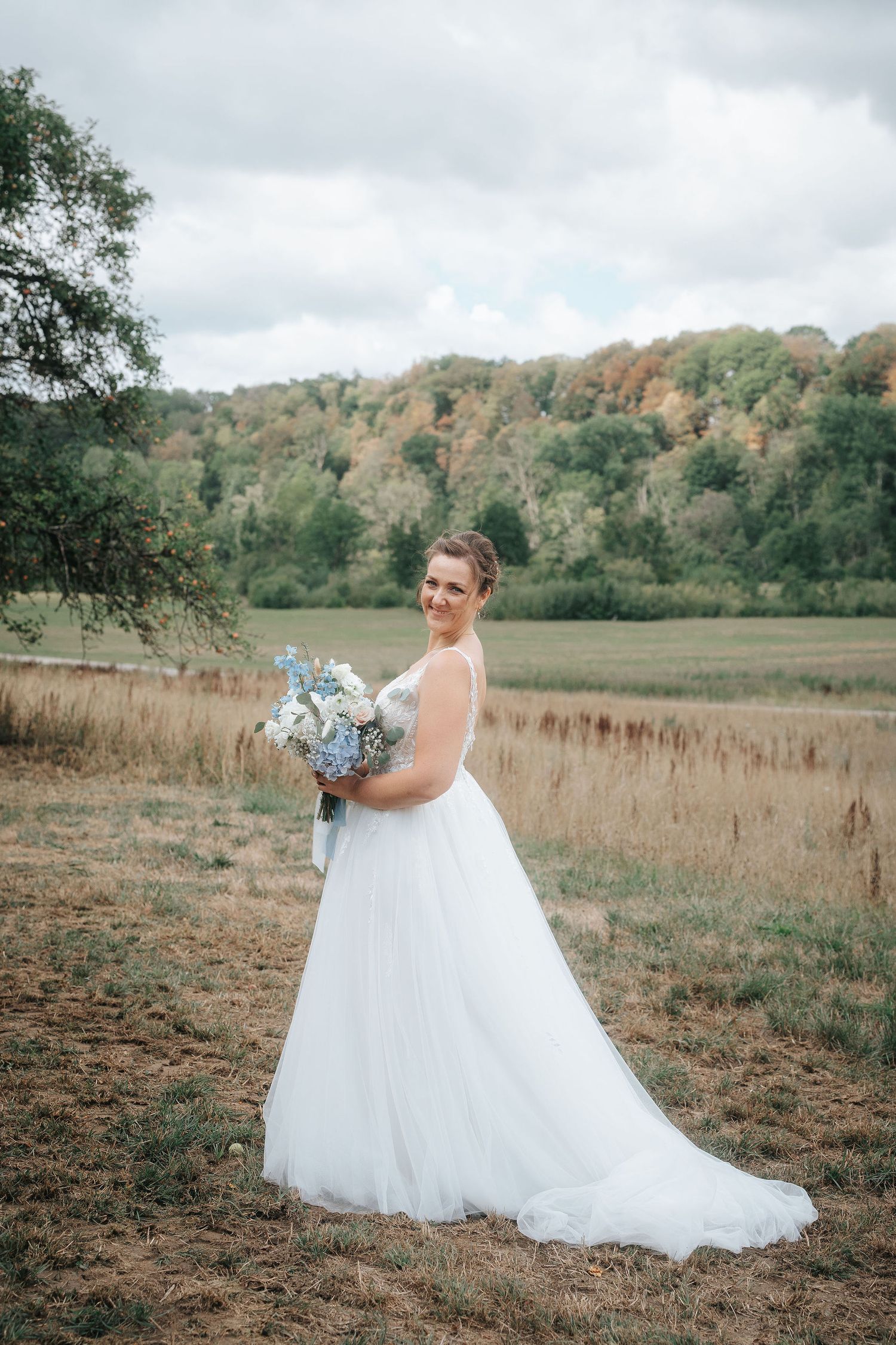 Bride in a flowing white tulle wedding gown holds a blue and white bouquet in a grassy field with autumn-colored hills behind her.