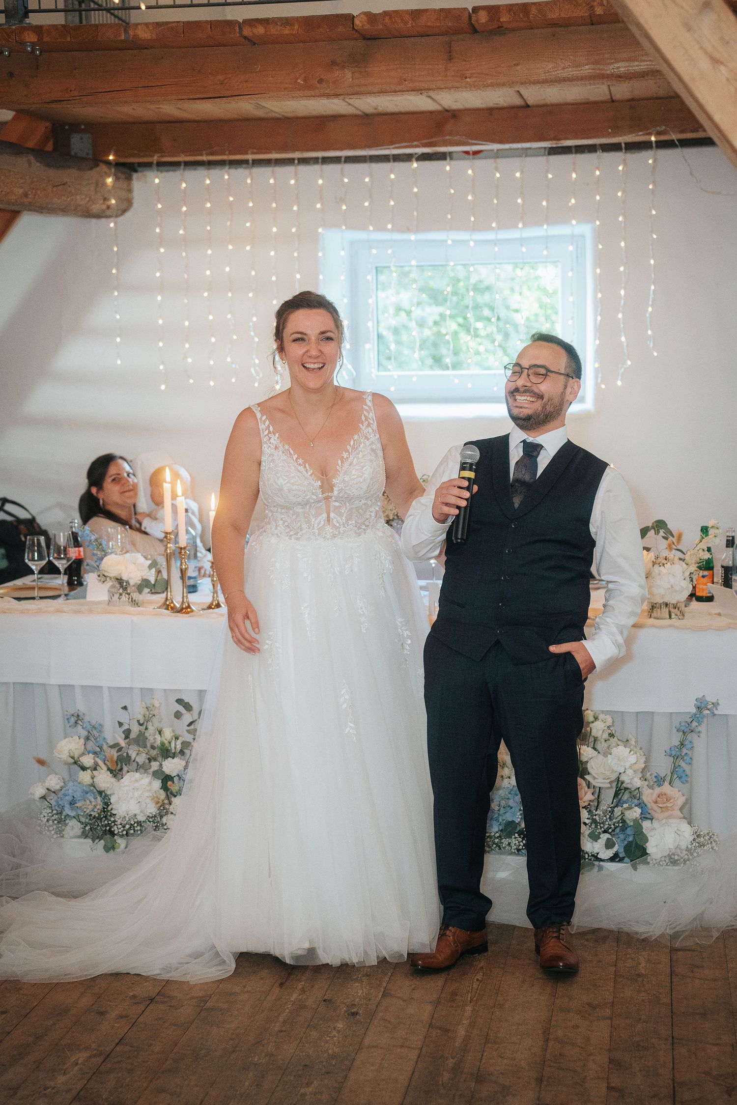 Smiling bride in a lace wedding gown stands beside a groom in a vest holding a microphone during a rustic barn wedding reception.