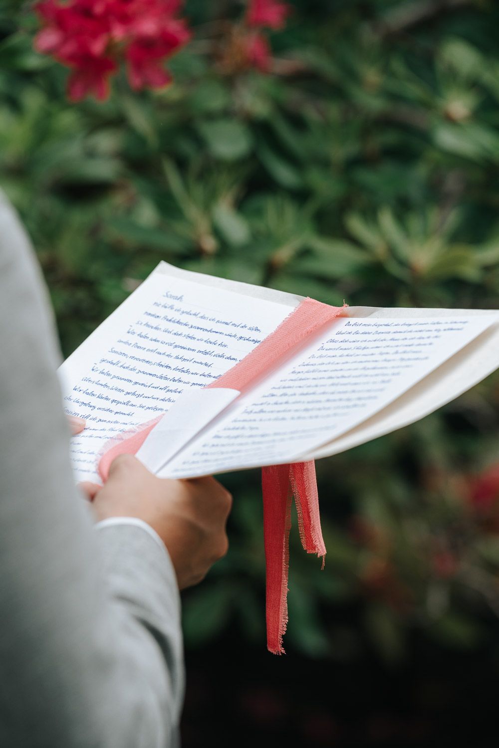 Someone holds an open book with a red bookmark against a background of blurred red flowers and green foliage.
