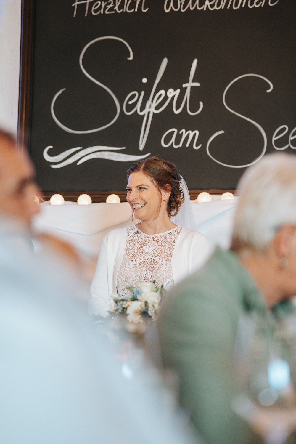 A person in a white lace wedding dress smiles at a reception with decorative lights and text written on a chalkboard.