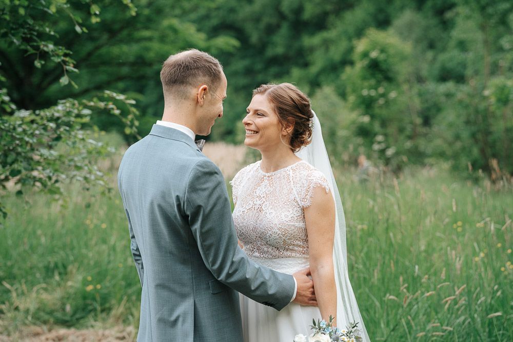 A romantic wedding couple stands together in a lush green meadow wearing a white lace gown and grey suit.