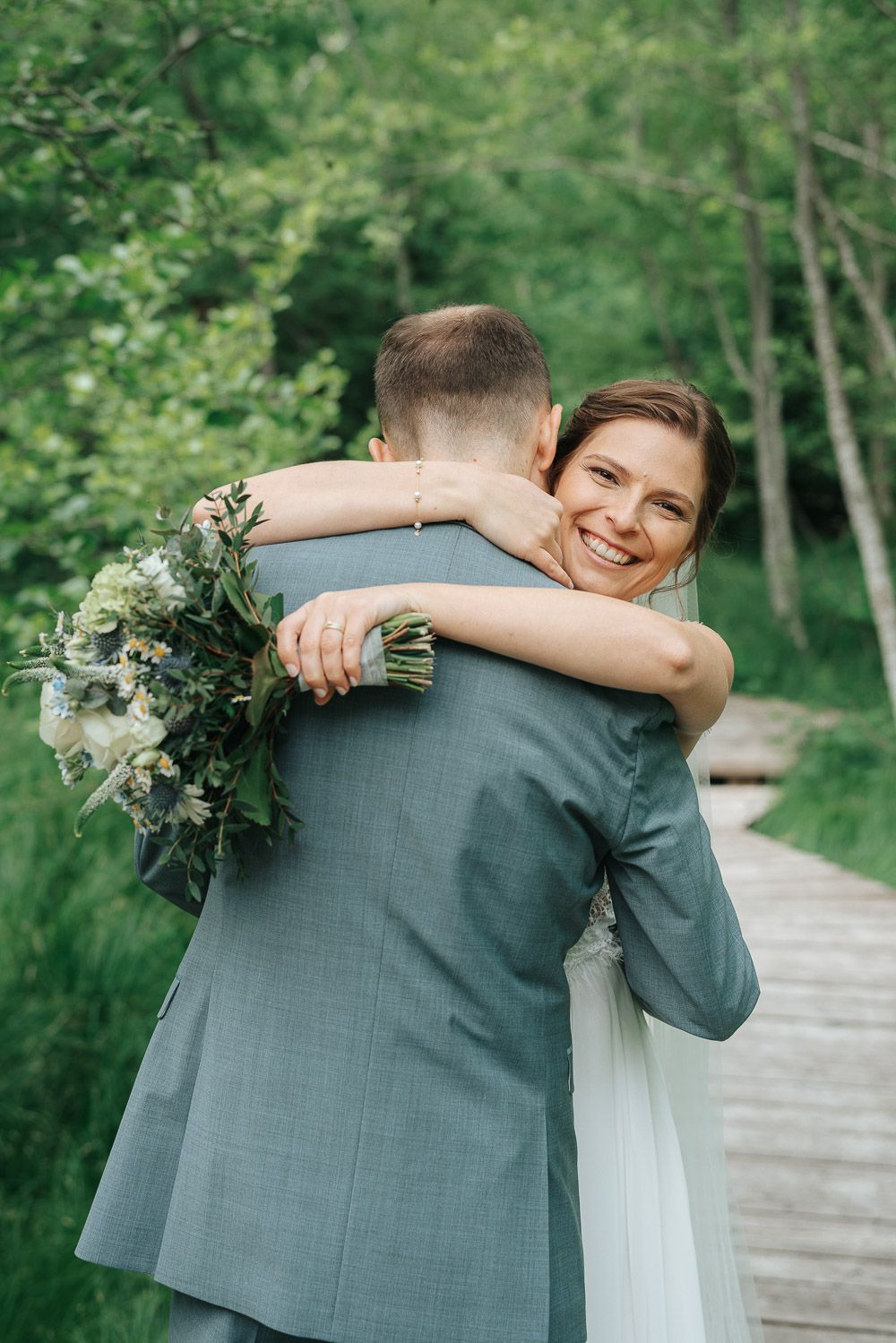 A joyful embrace between newlyweds on a wooden path surrounded by lush green forest, bride holding a natural bouquet.