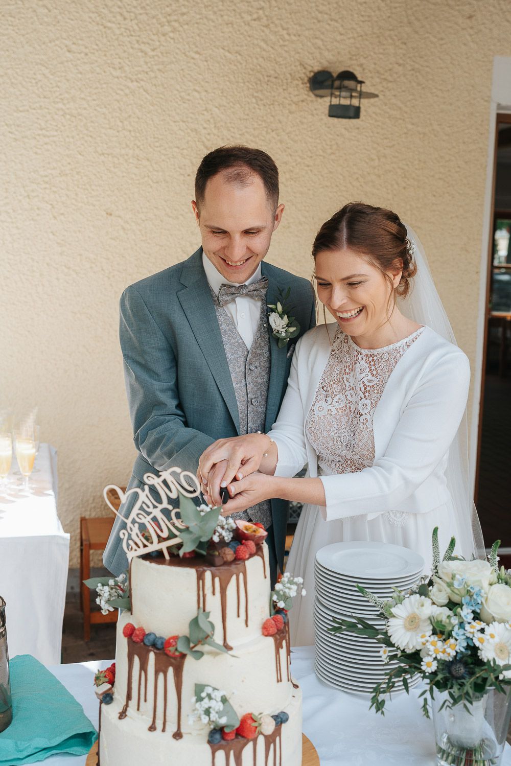 A couple sharing a joyful moment while cutting their white wedding cake decorated with chocolate drips and berries.