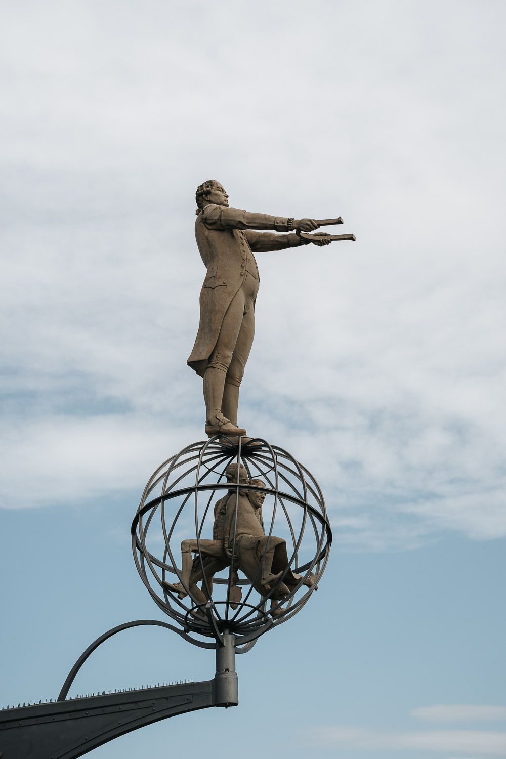 A statue figure in formal attire stands atop a spherical wire frame structure pointing outward against a cloudy sky.