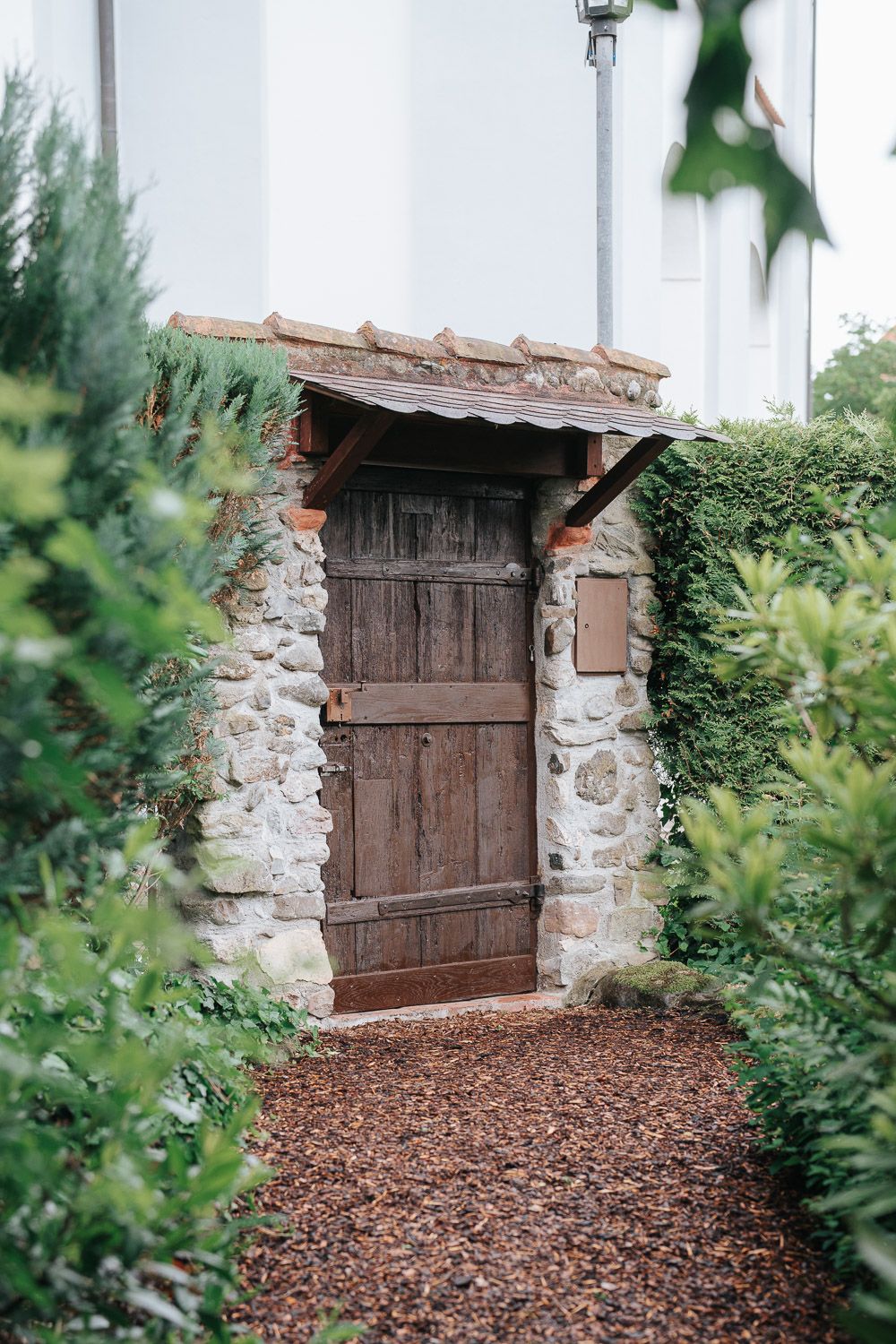 A rustic wooden door set in a stone wall with mulch path and green foliage creates an enchanting garden entrance.