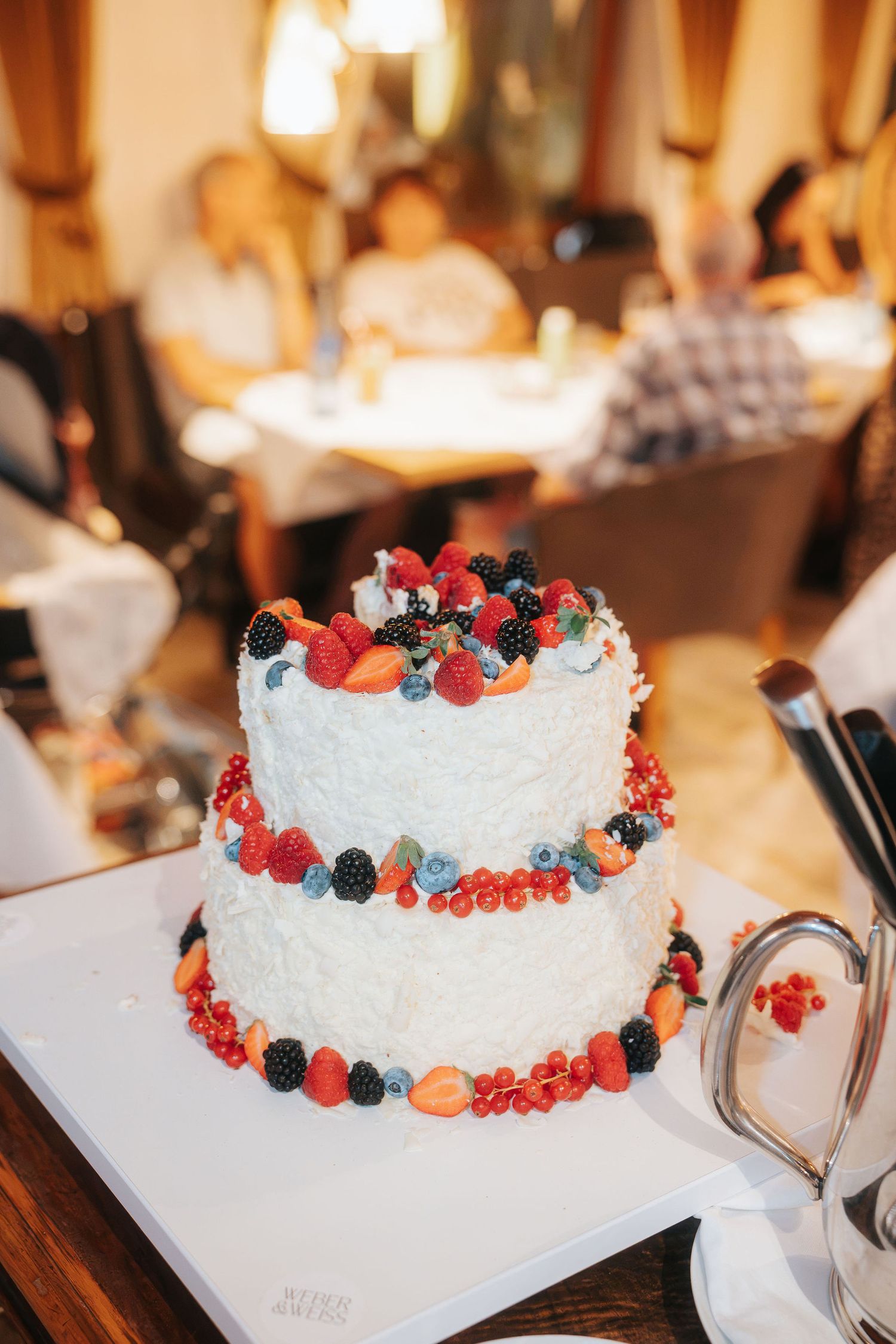 Two-tier white frosted cake decorated with fresh strawberries, blackberries, blueberries and raspberries on a white tray in a restaurant.