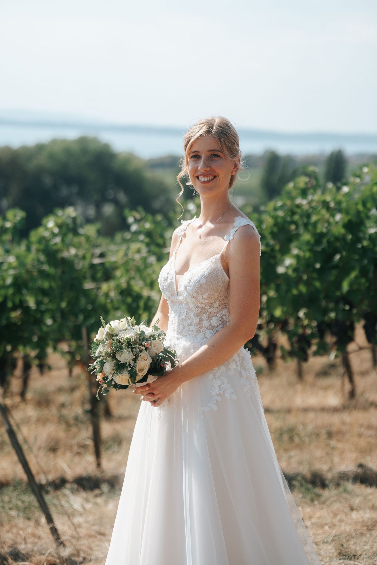 Smiling bride in a lace wedding gown holding a white floral bouquet in a sunny vineyard with a lake in the background.