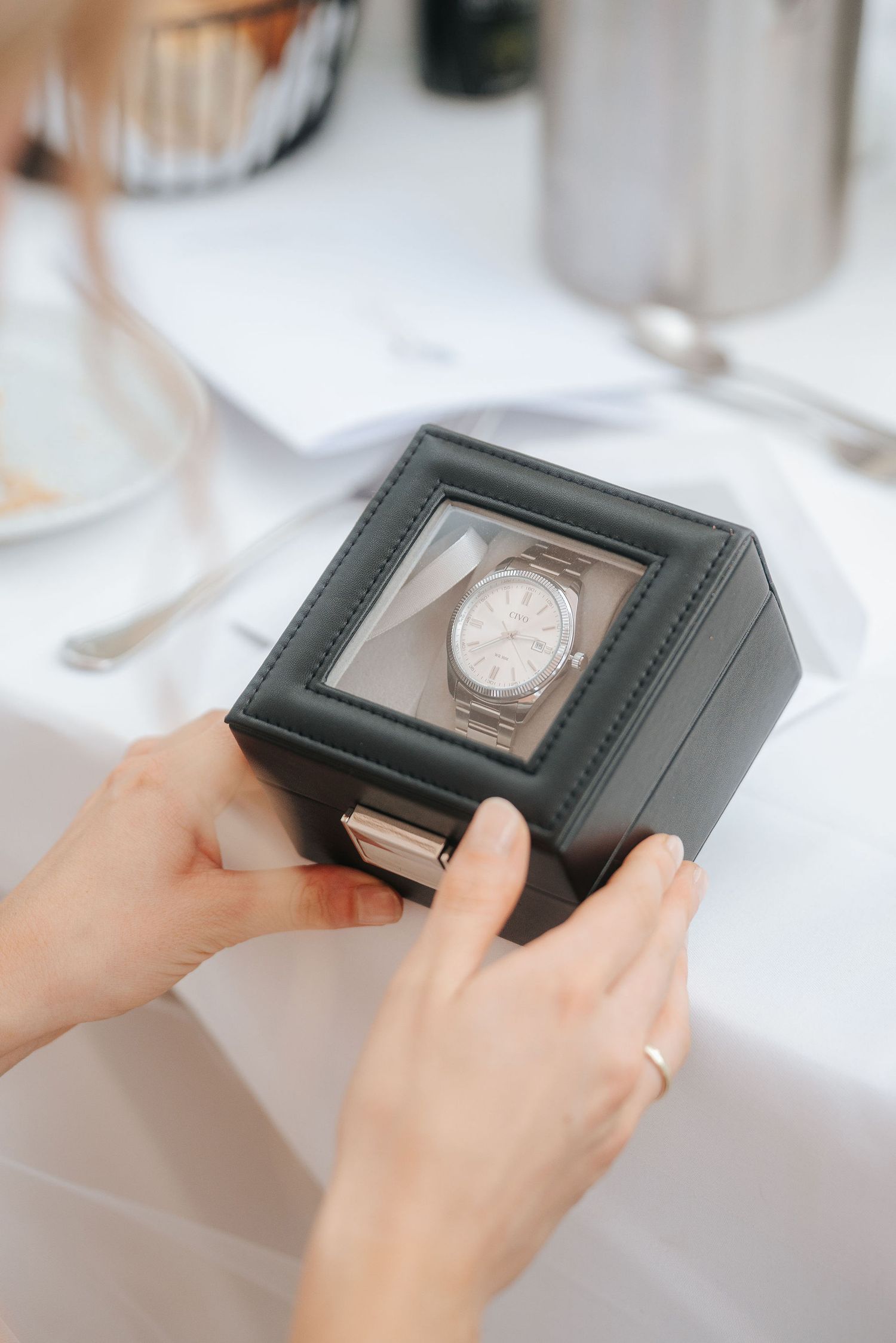 Hands holding a black leather watch box with a clear lid showing a silver wristwatch, set on a white table with cutlery nearby.