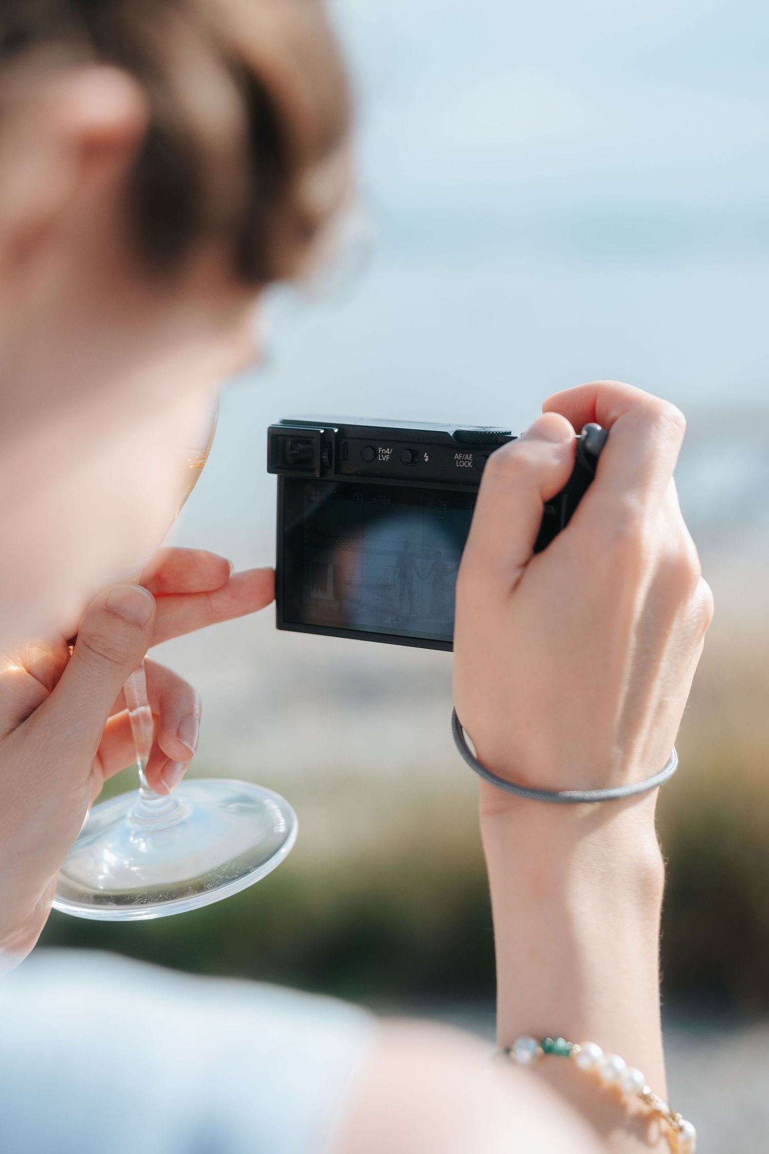 Woman holding a compact digital camera in one hand and a wine glass in the other while taking a photo outdoors on a sunny day.