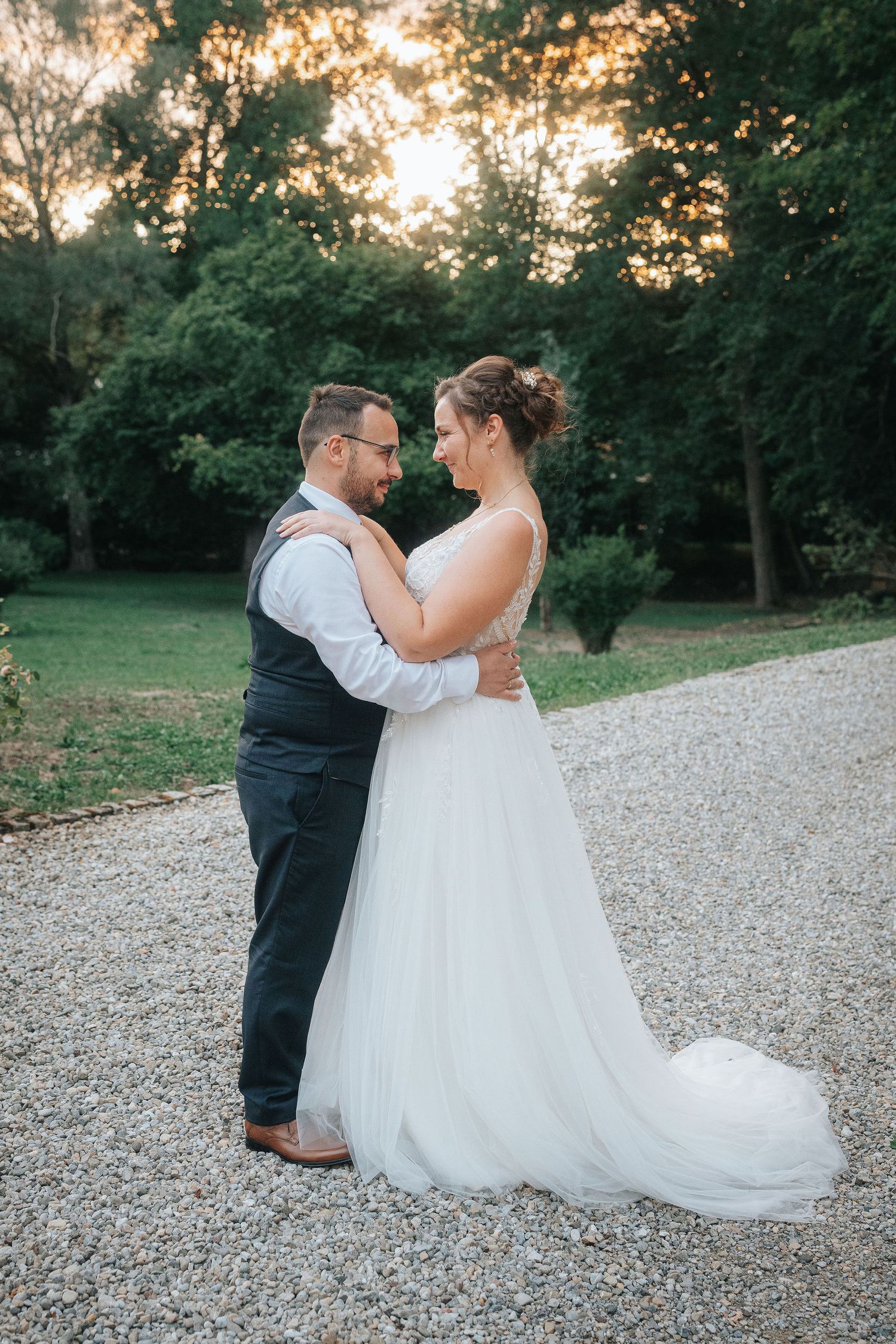 Bride in a flowing white gown and groom in a vest embrace on a gravel path at sunset, surrounded by green trees.