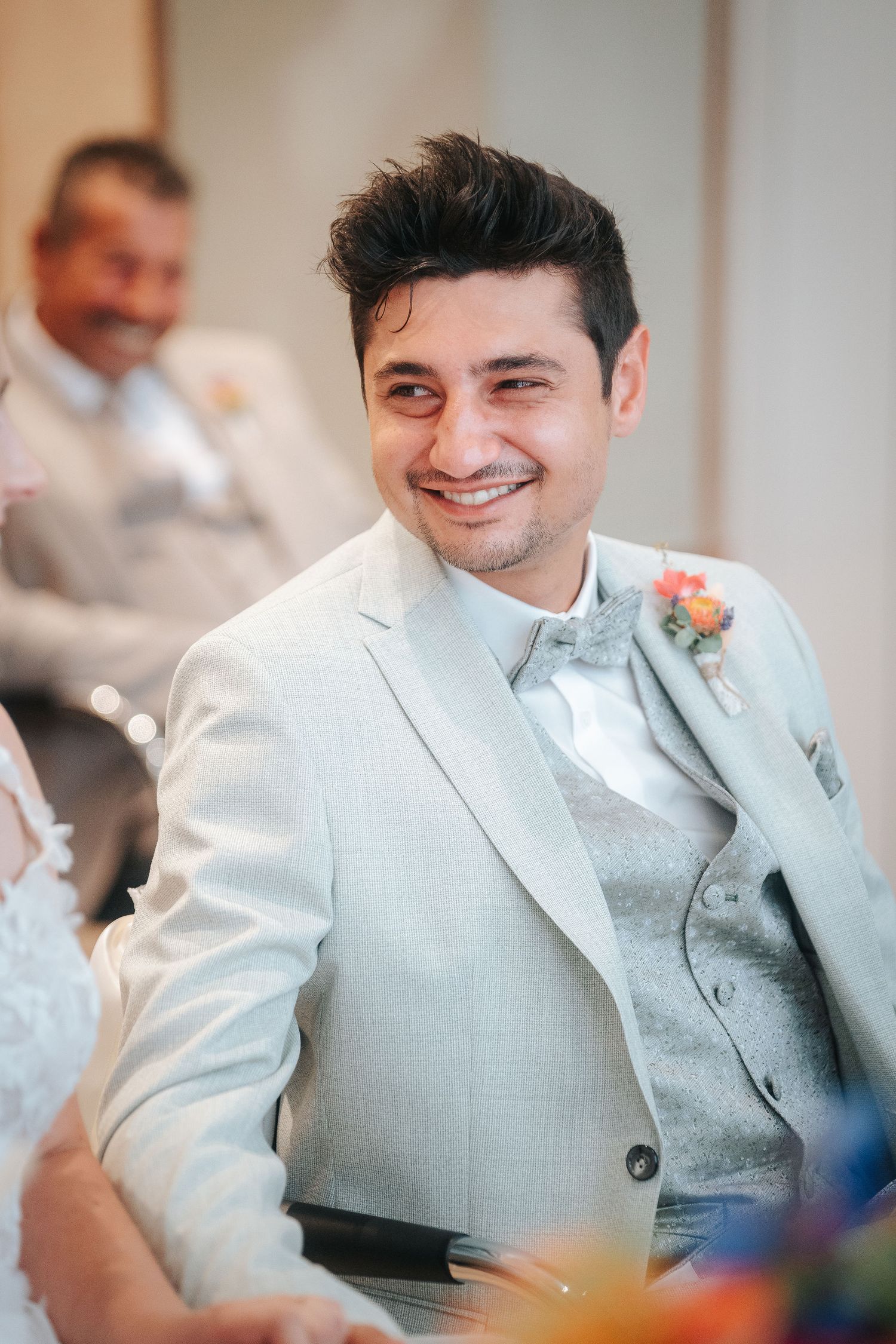 Smiling groom in a light gray suit with a floral boutonniere sits at a wedding ceremony while a guest looks on behind him.